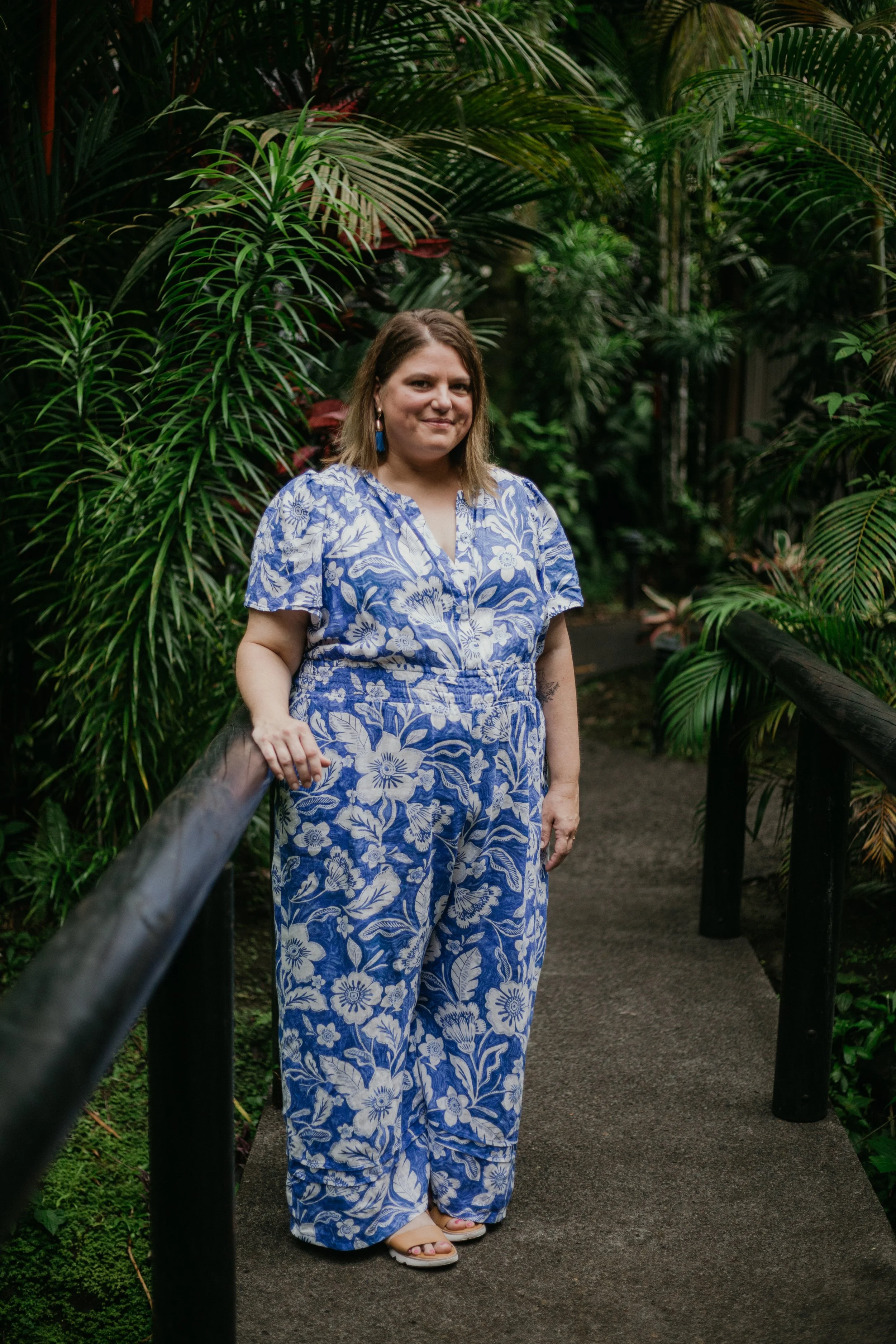 Woman standing on a small wooden pathway, surrounded by lush green tropical plants, wearing a matching blue and white floral patterned outfit and open-toed sandals.