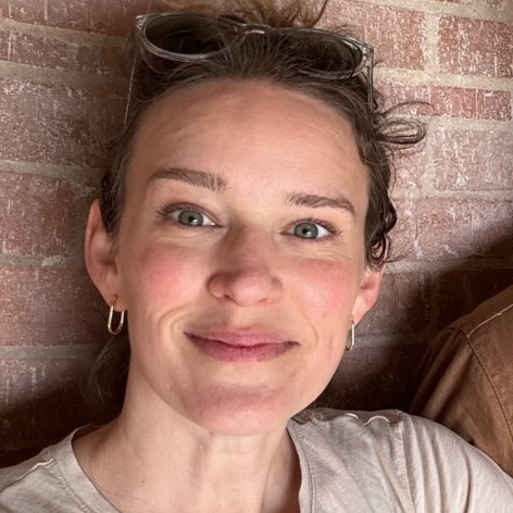 Person smiling indoors against a brick wall, wearing sunglasses on top of their head and a casual top.