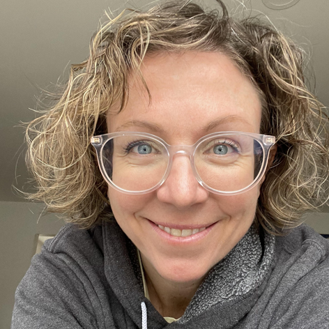 Person with curly hair and glasses smiling, wearing a gray hoodie.