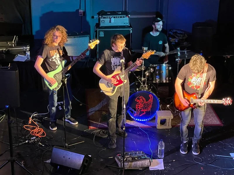 Band performing on stage with guitars and drums in a live music setting.