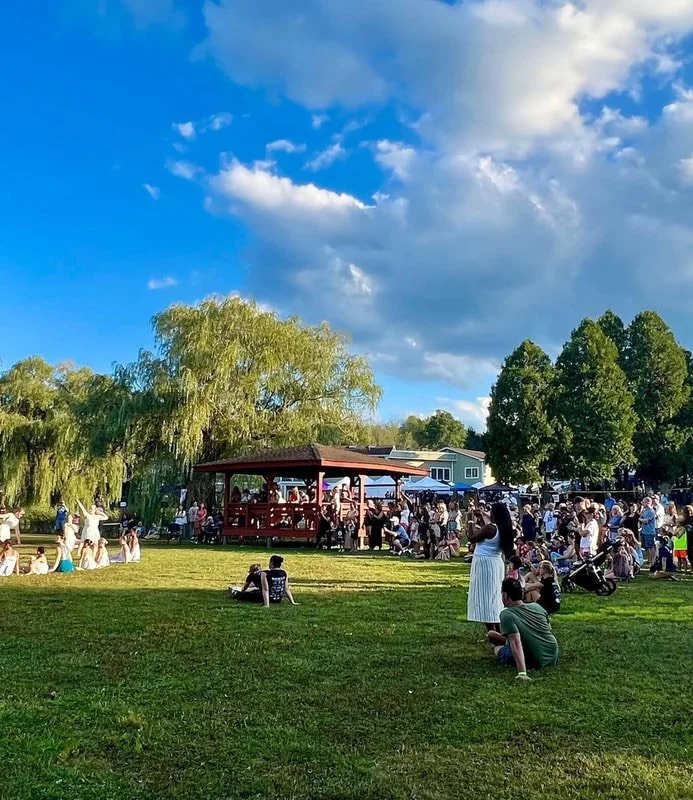 Outdoor event with people gathered around a gazebo on a sunny day, surrounded by trees and grass.