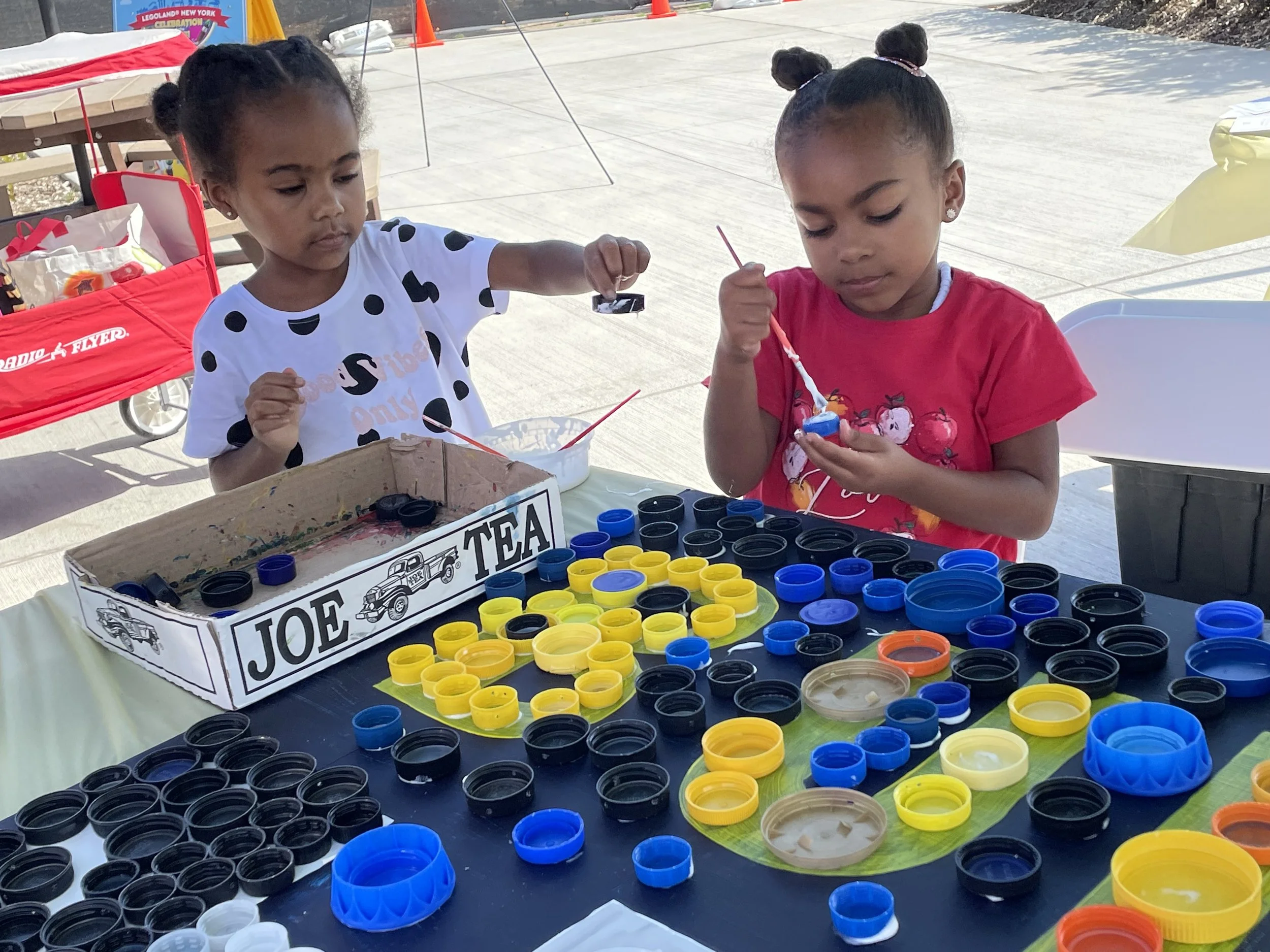 Two children painting plastic caps at a table outdoors, engaging in a creative activity.
