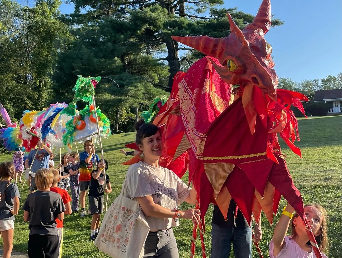 Outdoor event with people gathered around a gazebo on a sunny day, surrounded by trees and grass.