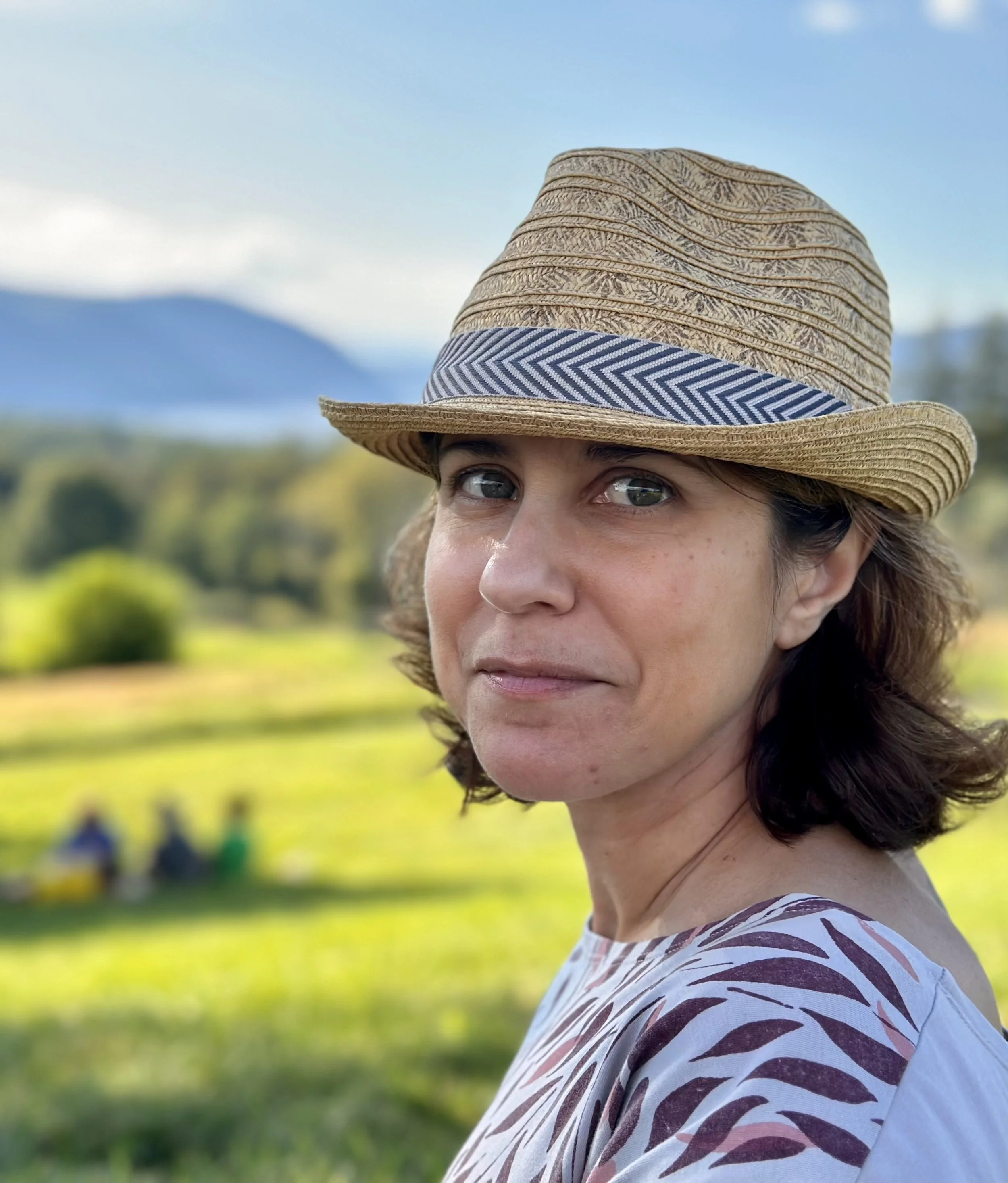 Woman wearing a straw hat in a park setting with mountains in the background.