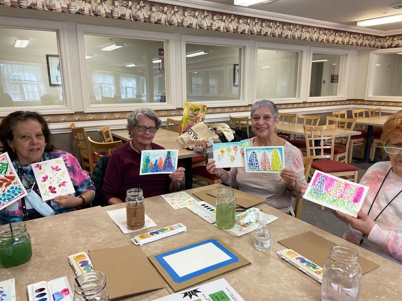 A group of elderly women sitting at a table in a craft room, each holding a watercolor painting with colorful designs. Art supplies, like watercolor palettes and jars, are on the table. The room has wooden chairs, mirrors, and windows.