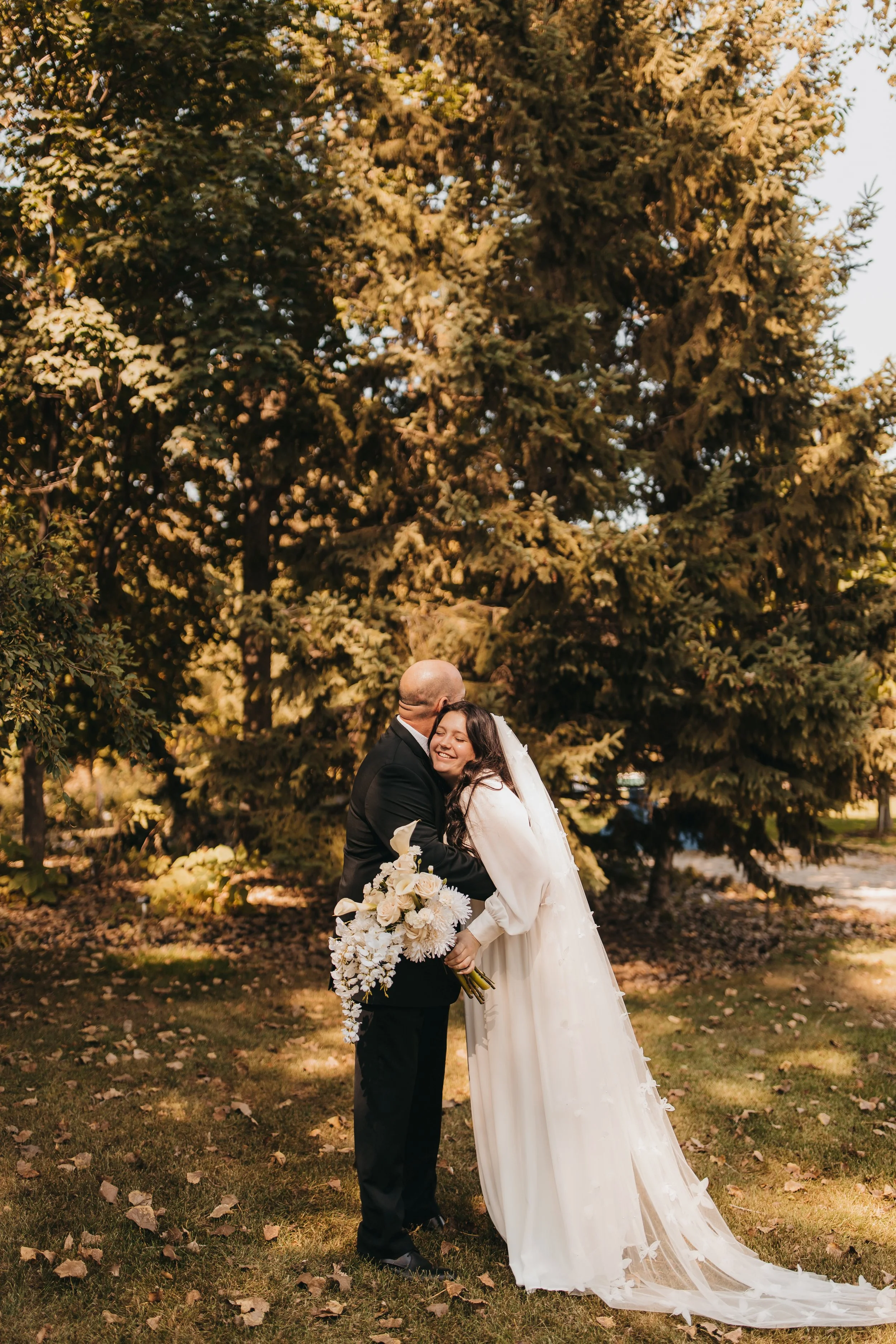 A bride and groom hugging outdoors with large evergreen trees in the background, the bride wearing a white wedding dress and holding a bouquet of white flowers.