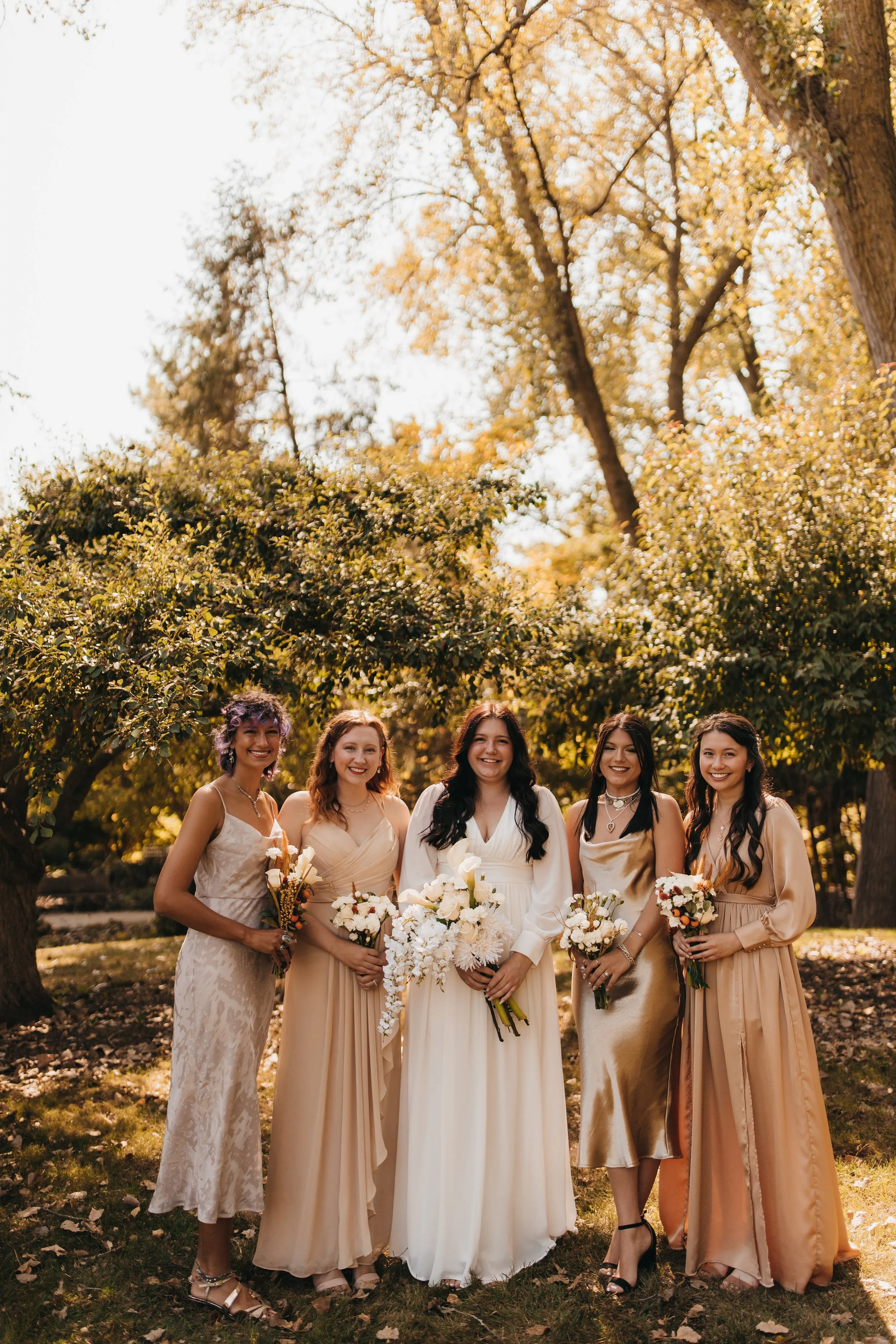 Five women in dresses holding bouquets, standing outdoors among trees on a sunny day, celebrating a special occasion.
