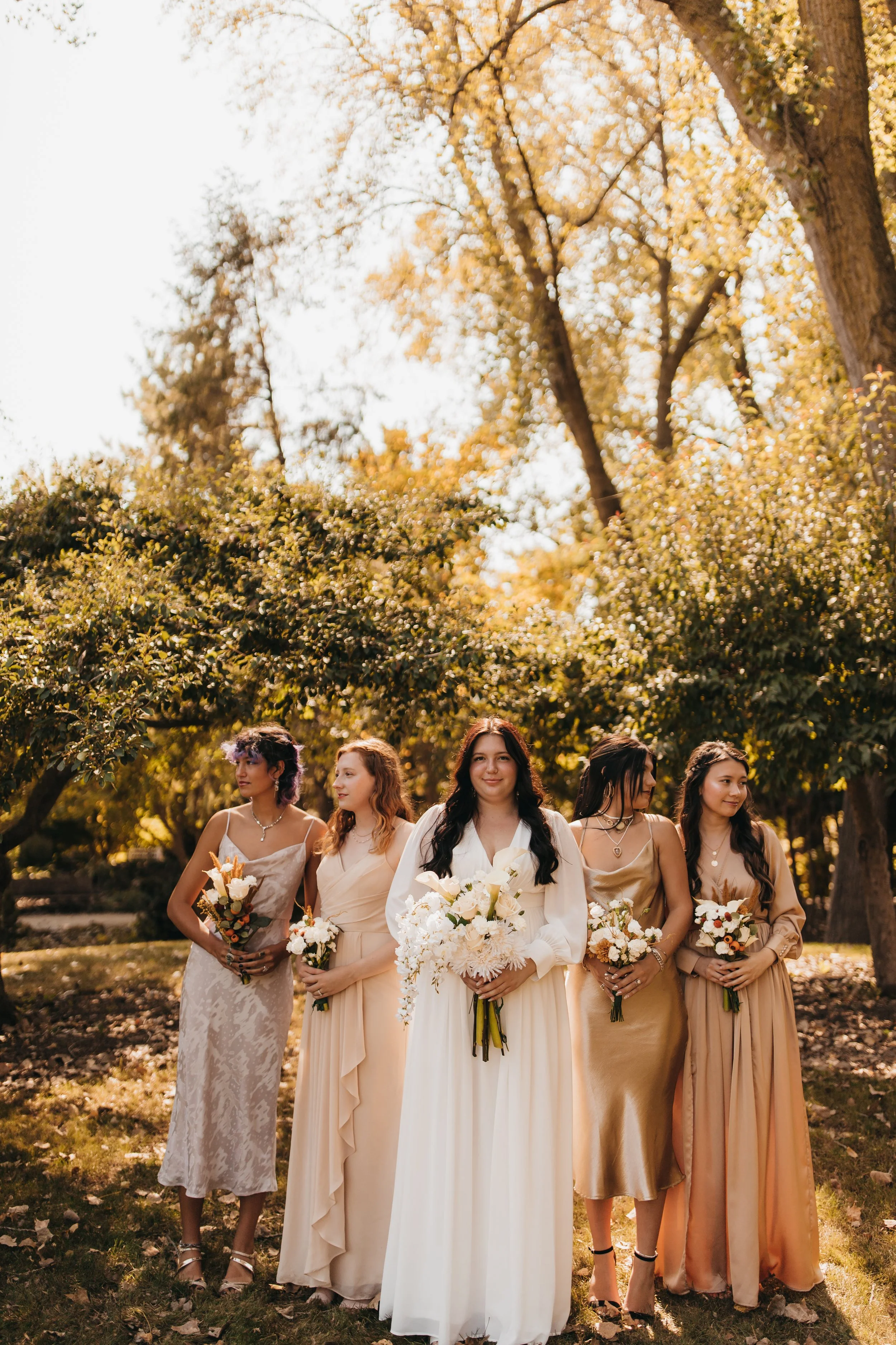 Group of five women dressed in elegant gowns holding bouquets, standing outdoors under trees with autumn foliage