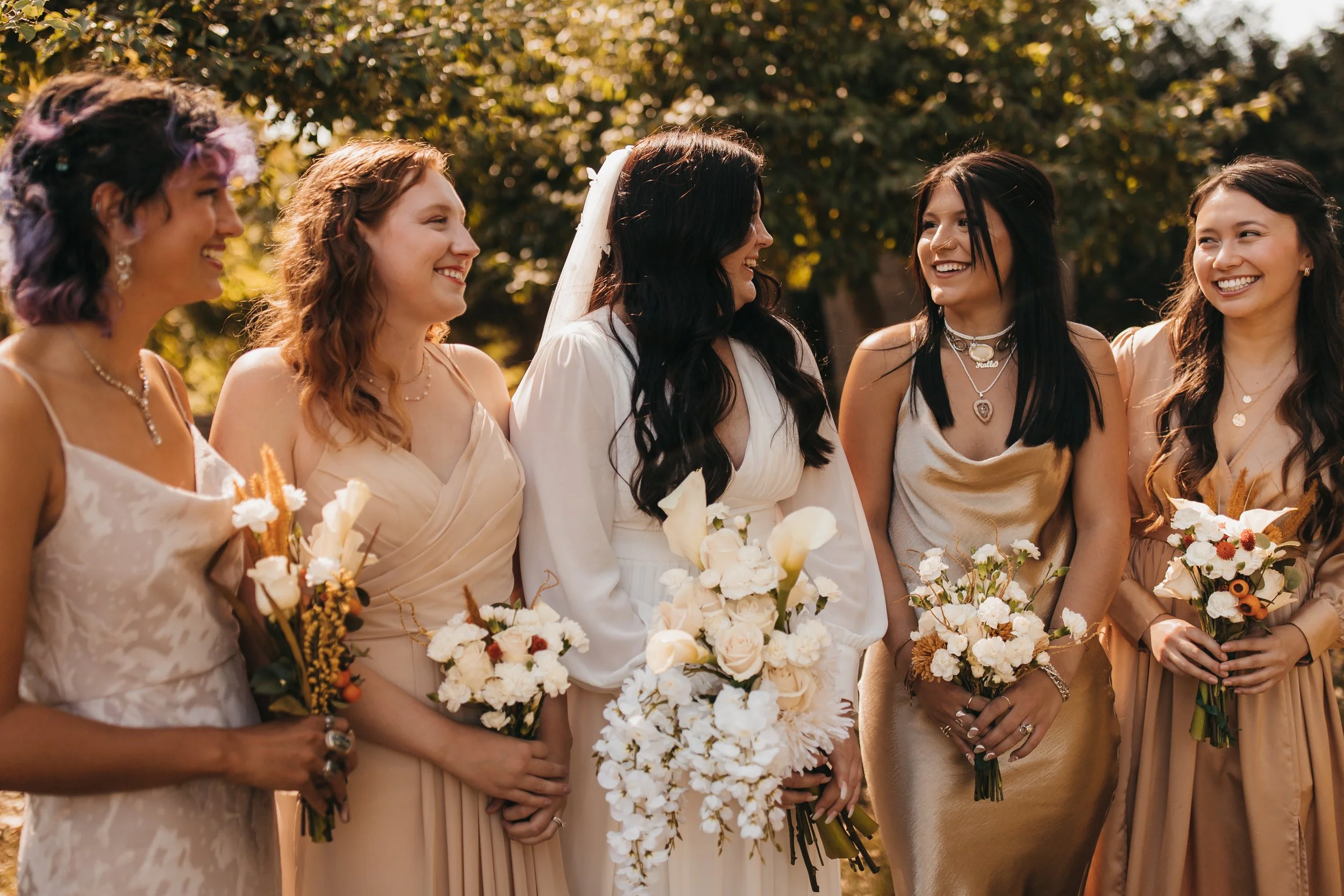Group of women in light-colored dresses holding bouquets at a wedding outdoors, smiling and chatting.