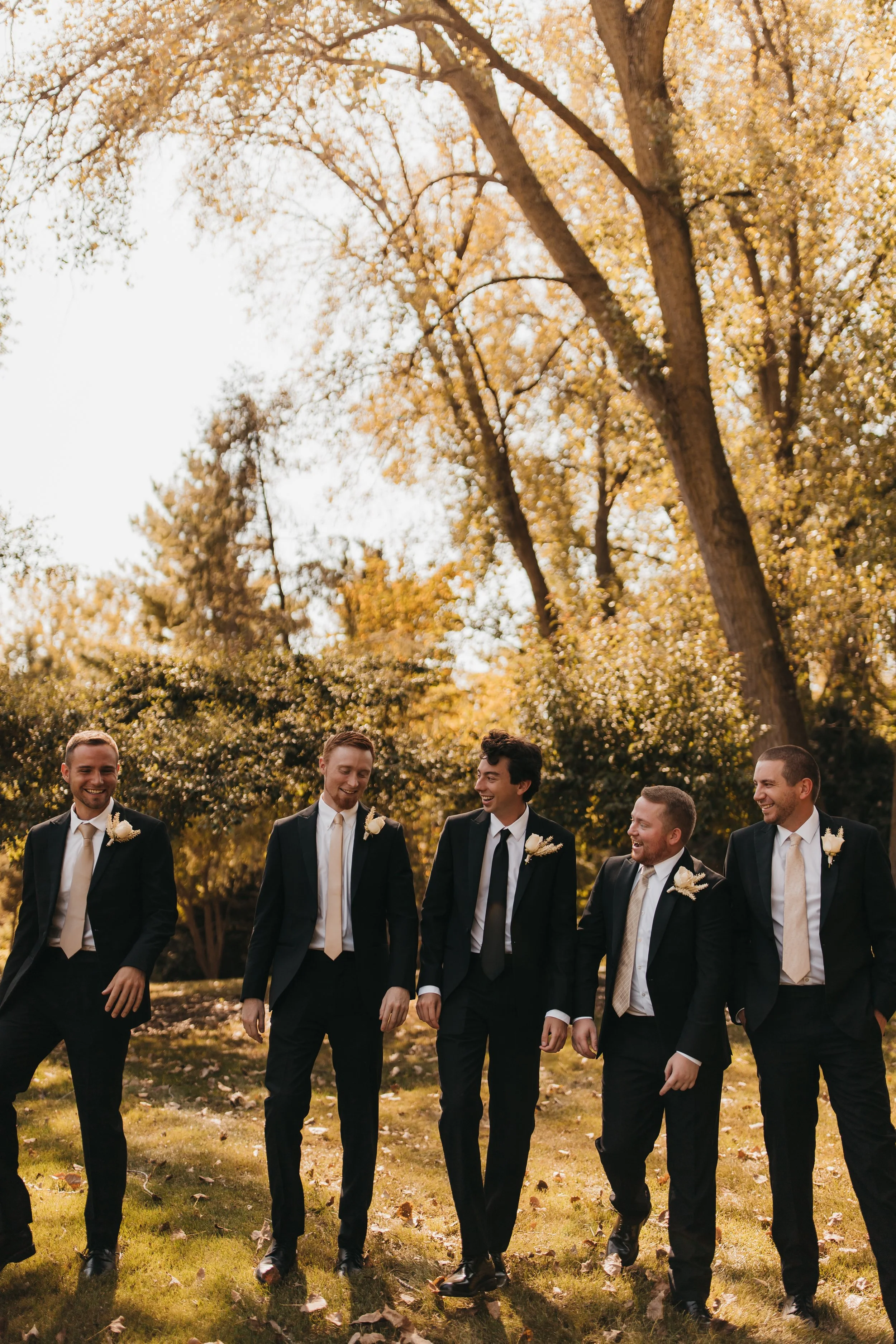 Group of five men dressed in suits and ties walking and smiling outdoors in a park with autumn foliage.