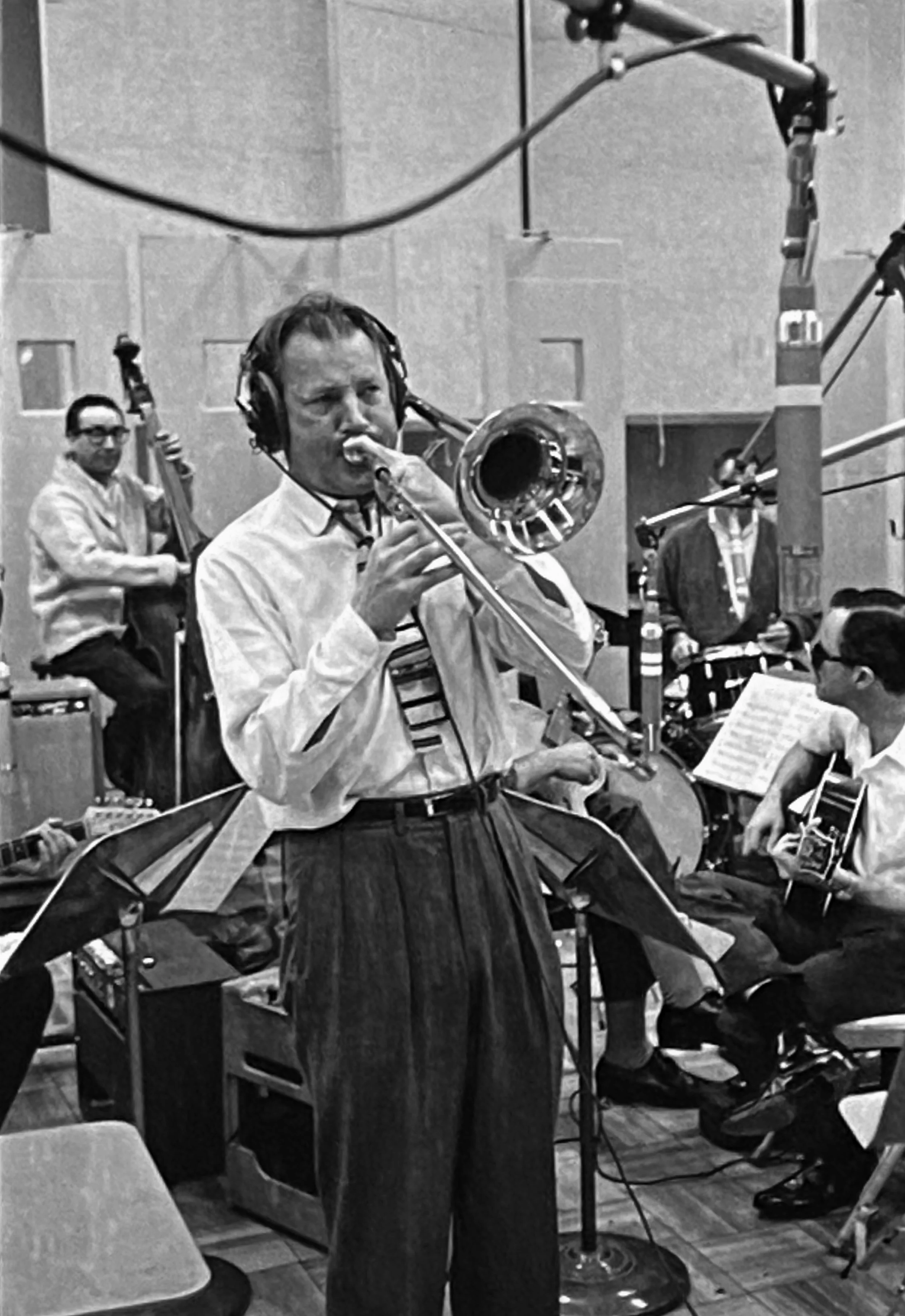 Musical ensemble recording session with a man playing the trombone in the foreground, a bassist and a guitarist in the background, in a recording studio.
