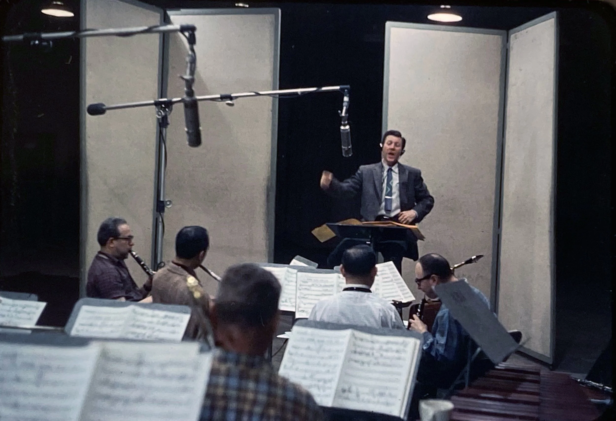 A group of musicians in a recording studio or rehearsal space, with a conductor or director standing on a platform, conducting or leading the musicians, who are playing wind instruments and reading sheet music.