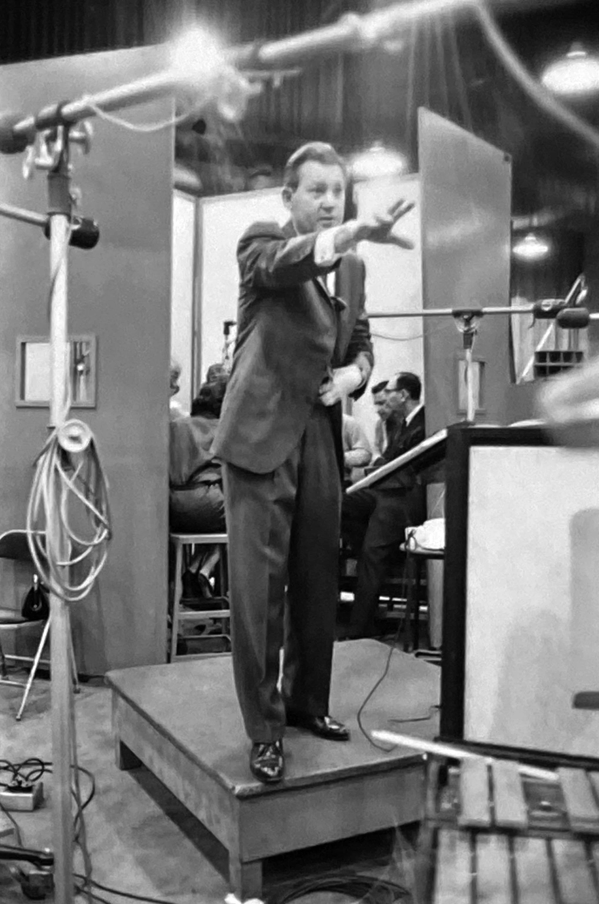 A man in a suit is standing on a small podium, gesturing with his right hand, in front of a microphone on a stand, during a recording session or interview in a studio.