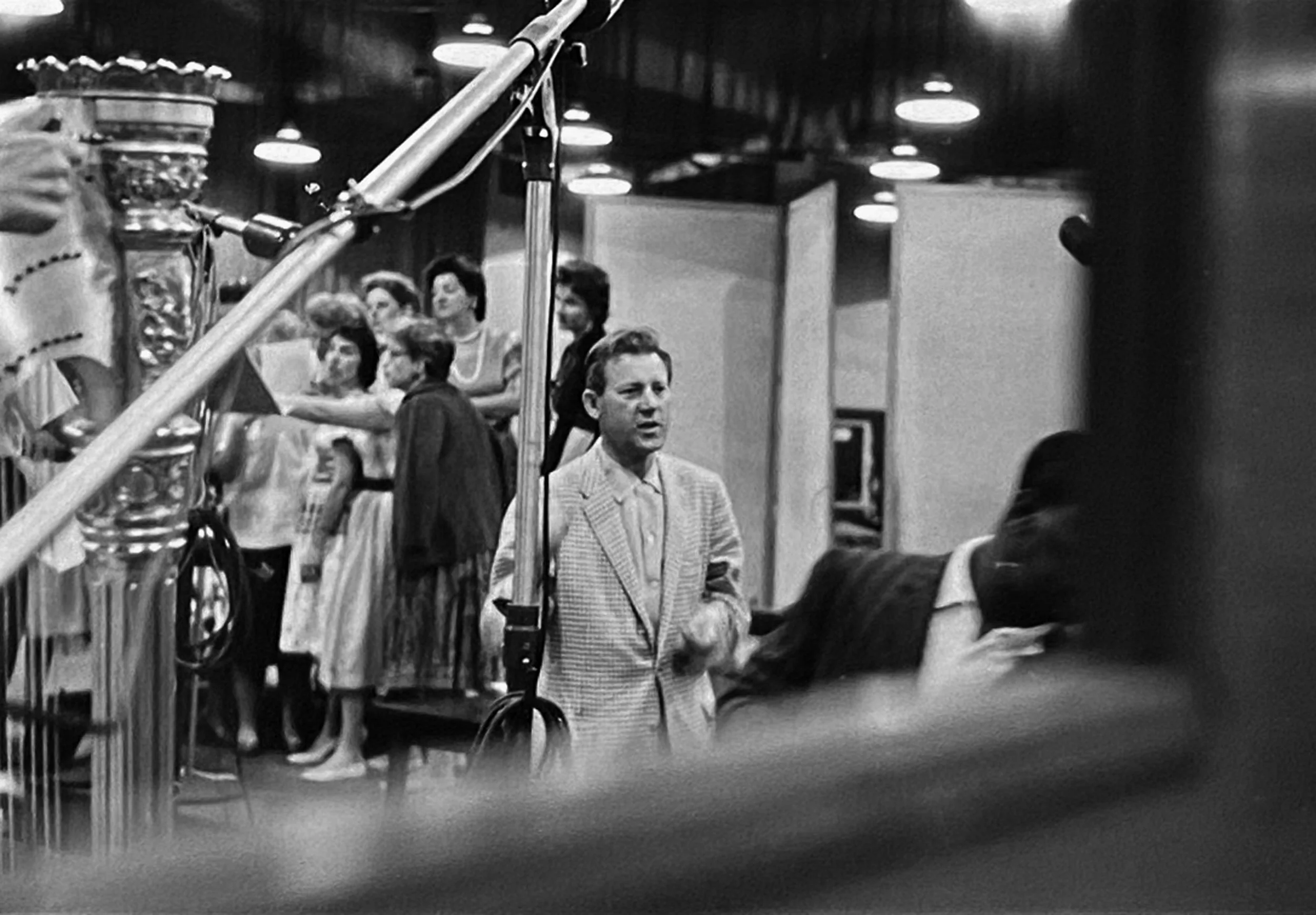 A black and white photo of a man in a suit speaking or singing in front of a group of women. The women are standing on a staircase, holding papers, and appear to be part of a choir or singing group. The setting looks like a stage or performance space