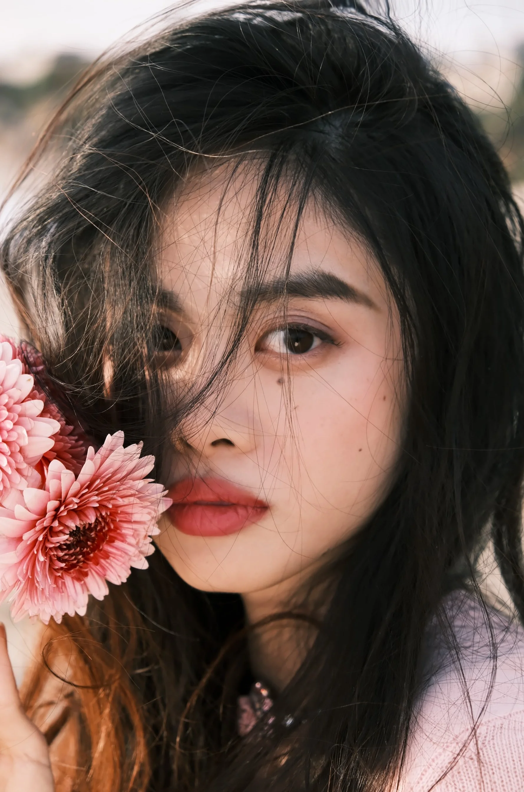Close-up portrait of a woman with long black hair holding a pink flower near her face, with some hair strands falling over her face, outdoors in soft lighting.