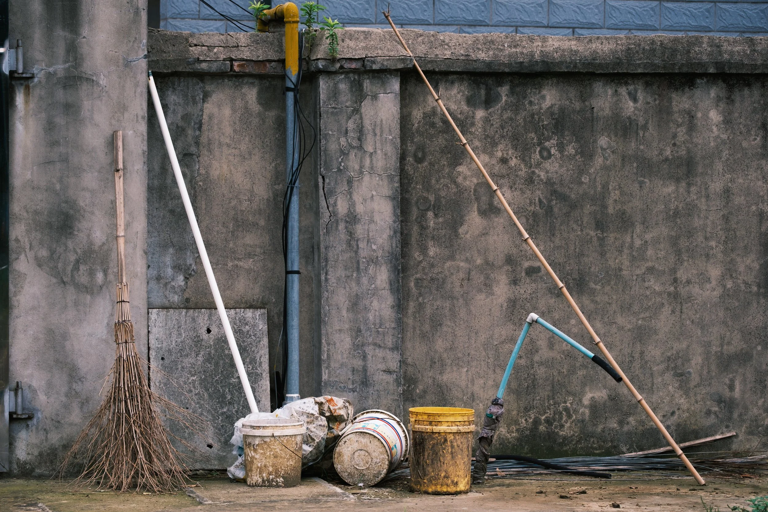 Cleaning supplies and tools against a concrete wall, including a broom, buckets, and poles.