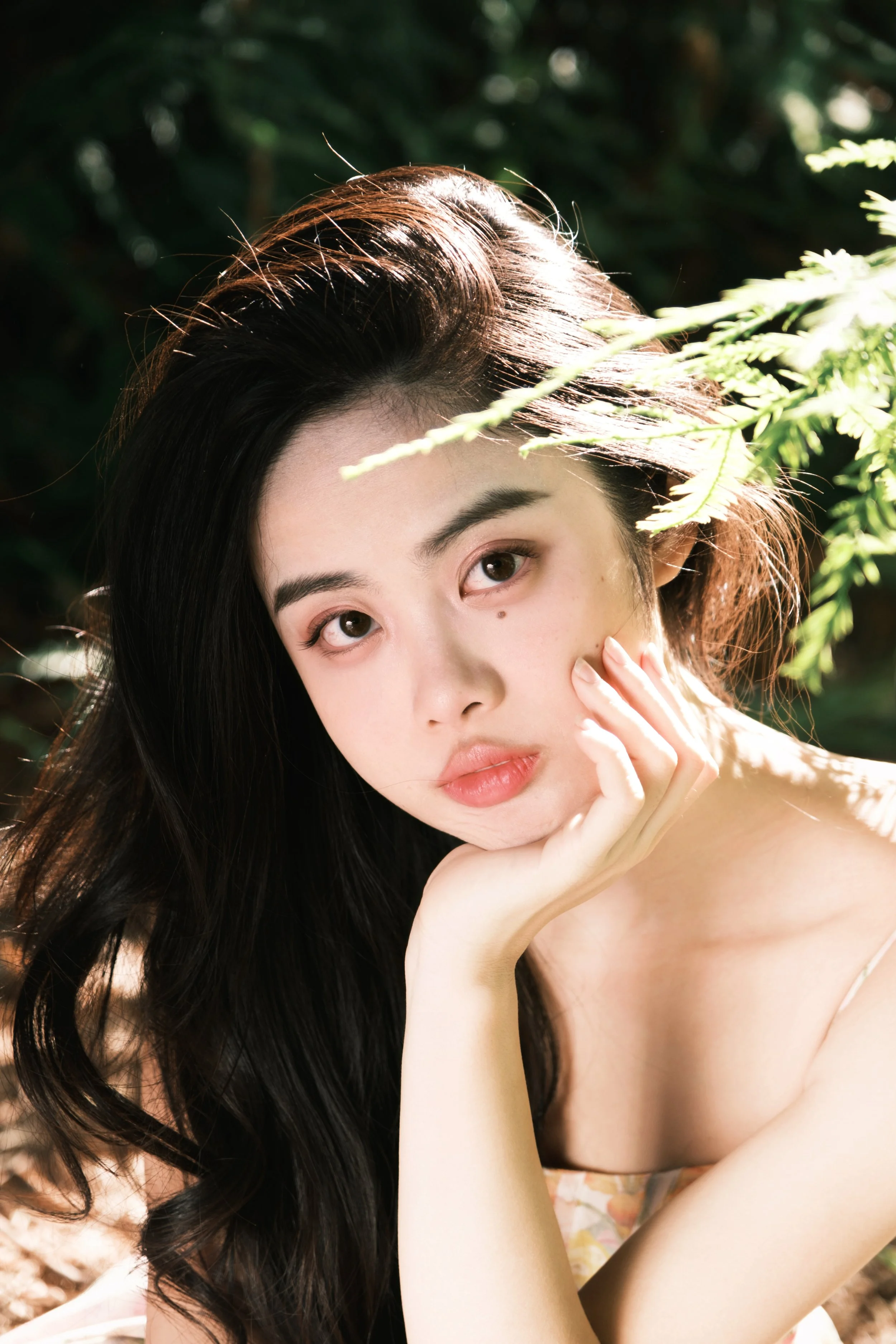 Young woman with long dark hair resting her face on her hand outdoors with green foliage in the background.