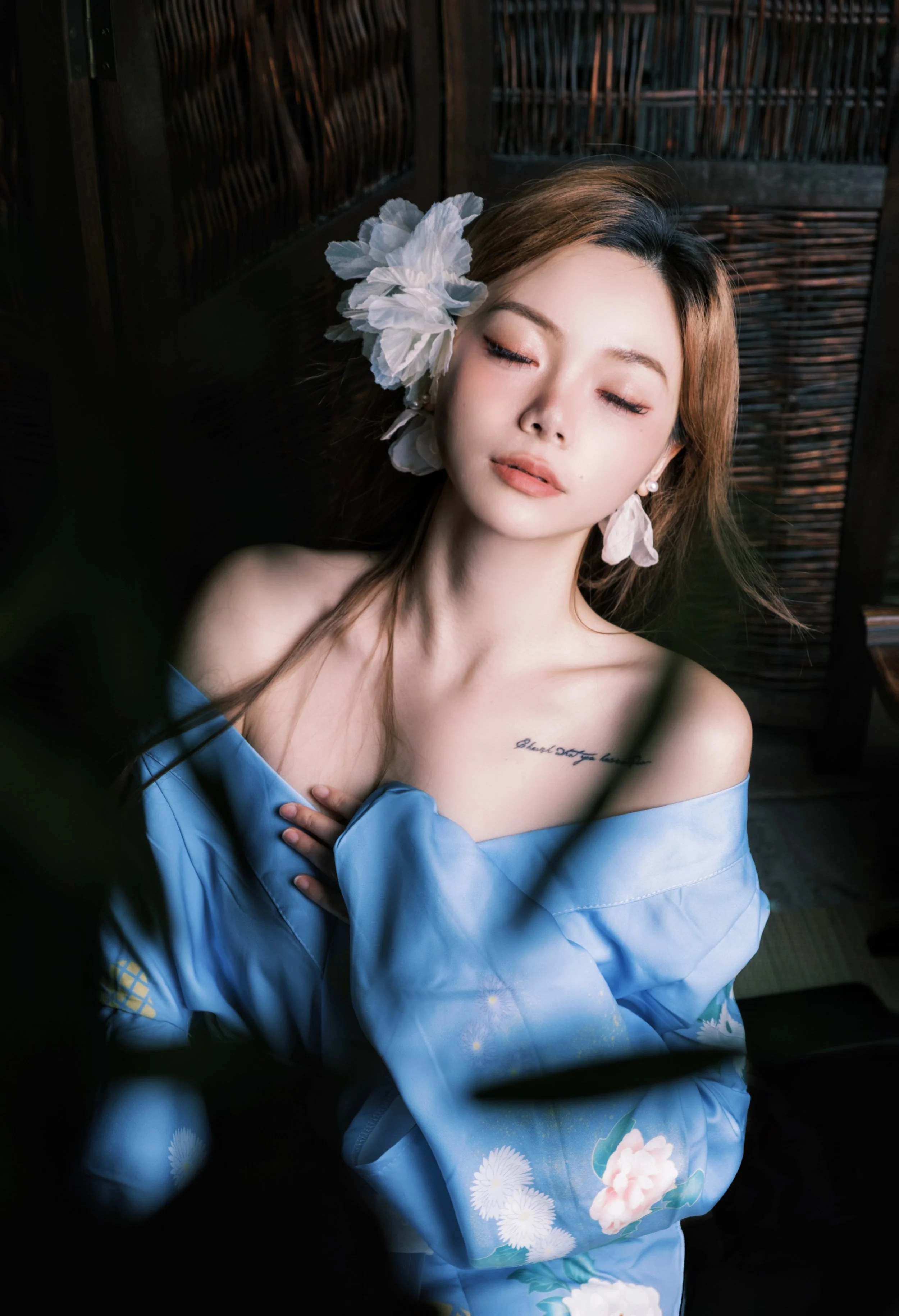 A woman with long brown hair and a white flower in hair, eyes closed, wearing a blue floral kimono, sitting indoors with wooden lattice background.