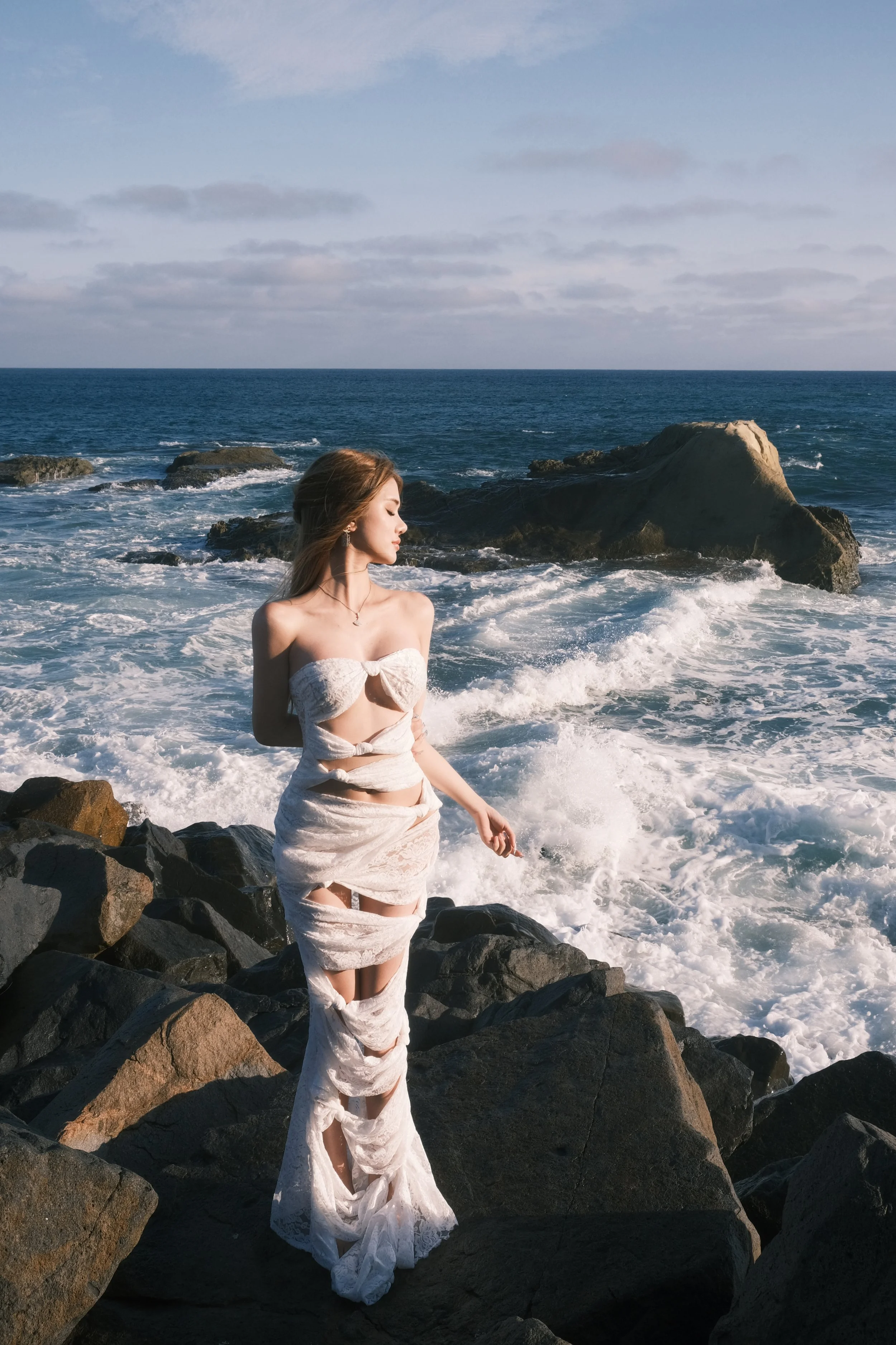 A woman in a torn, off-shoulder white dress standing on rocks at the beach with ocean waves crashing behind her and cloudy sky above.