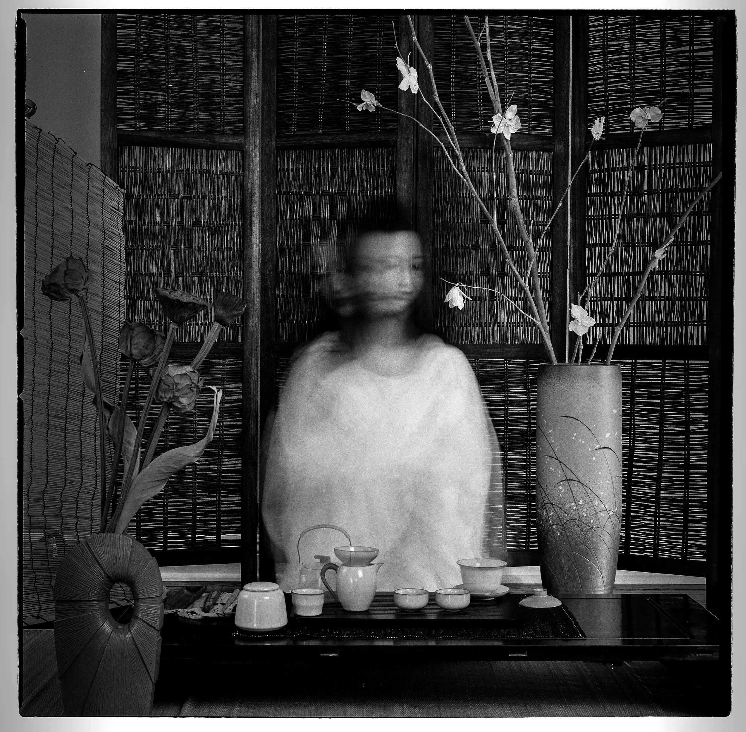 A blurred woman standing behind a traditional Japanese tea setup with teacups, teapot, and a jug on a tray, in front of two tall vases with flowers, with bamboo screens in the background.