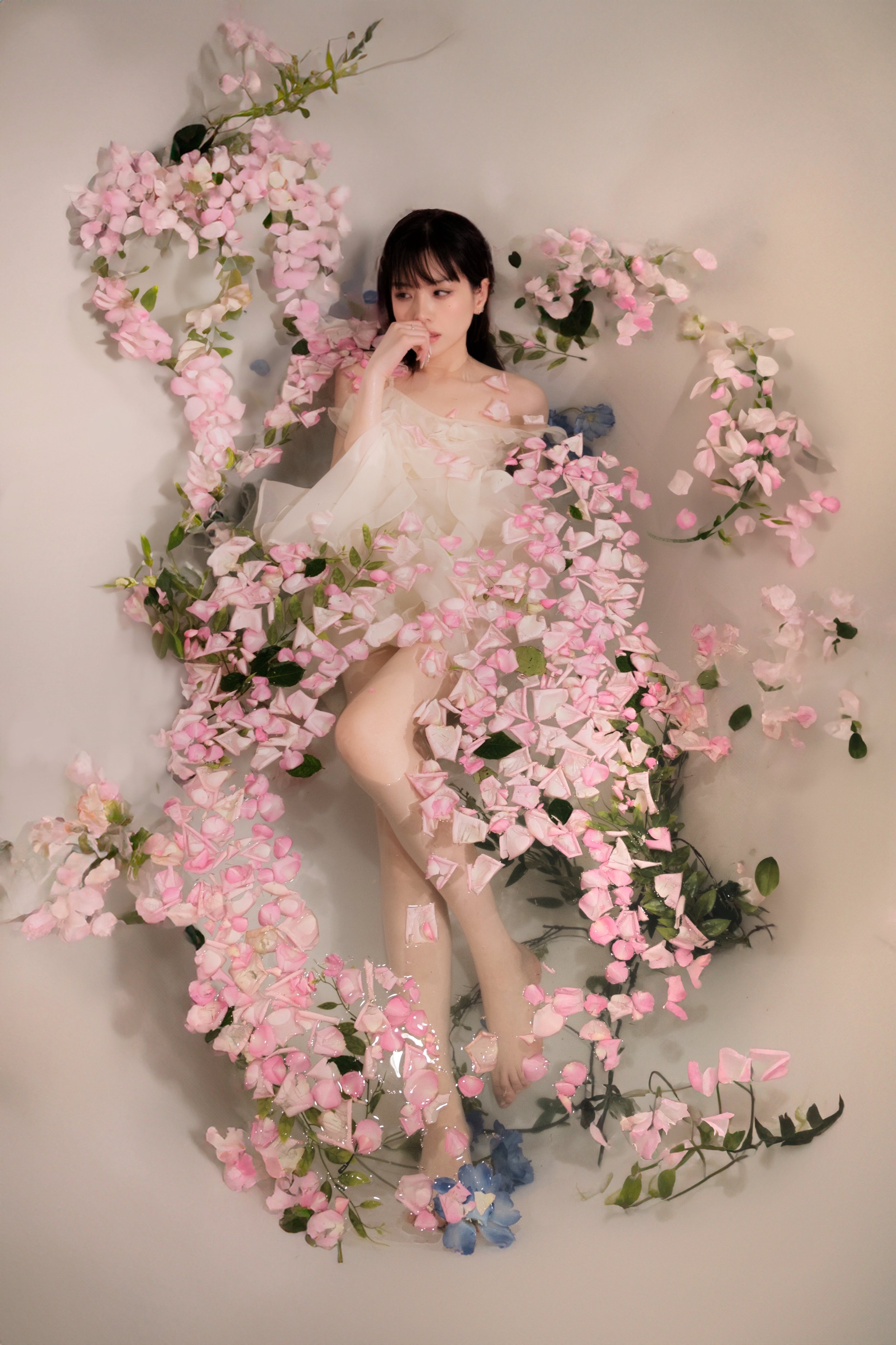 A woman lying among pink and blue flowers, with some petals on her face and body, against a plain light-colored background.