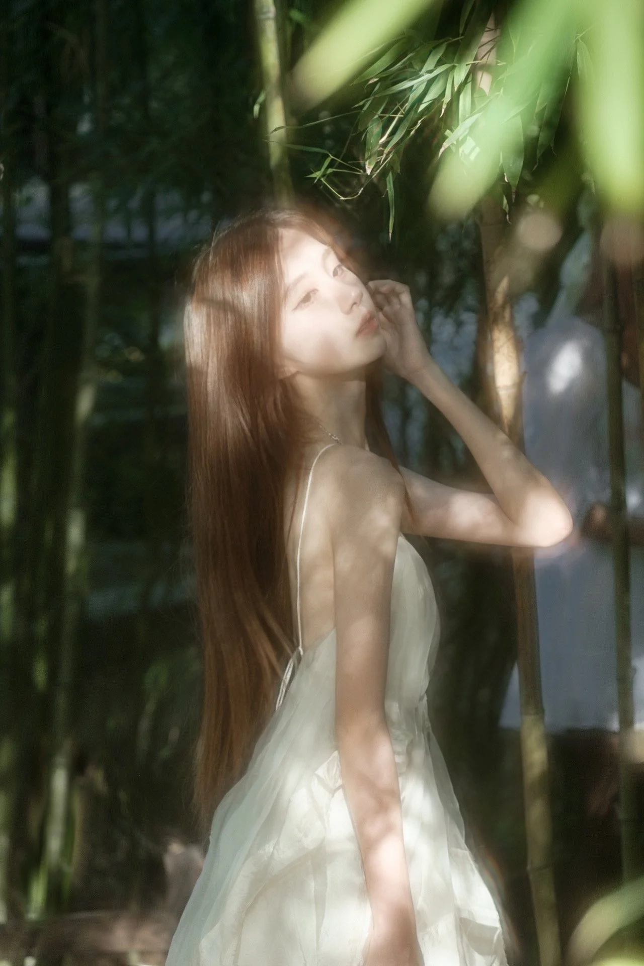 A woman with long brown hair wearing a light-colored dress standing behind a glass window with reflections, greenery, and trees visible in the background.