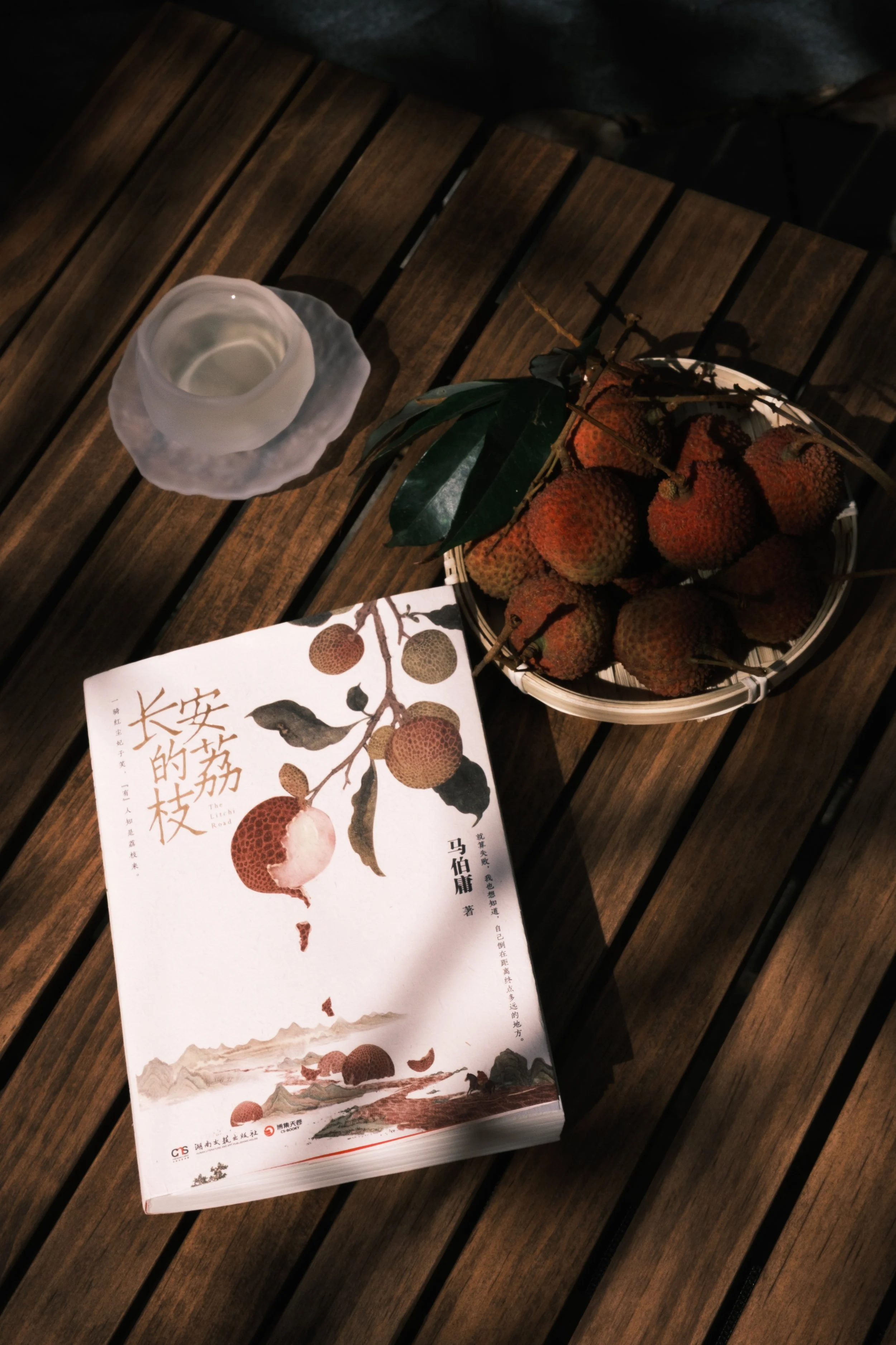 A wooden table holding a white book, a basket of lychee fruits, and a glass of clear liquid.