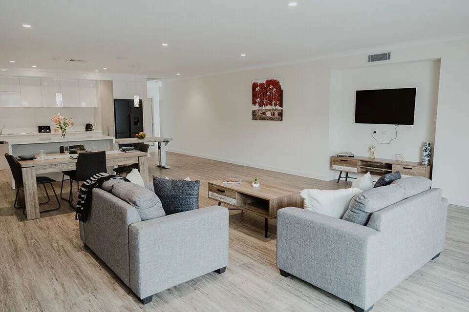 Modern open-plan living space with gray sofas, a wooden coffee table, dining area, and kitchen in the background.
