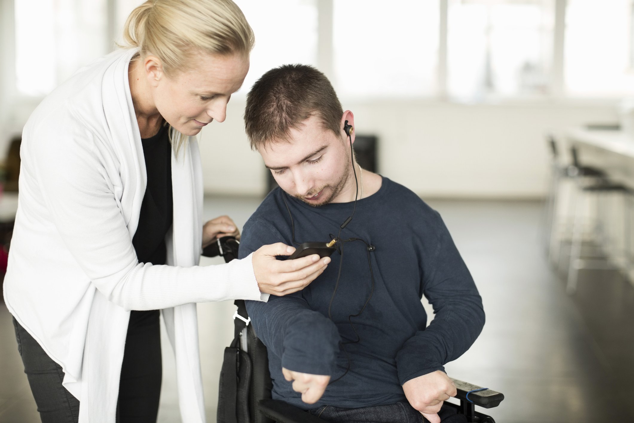 A woman assisting a man in a wheelchair, showing him something on a smartphone. The man is wearing headphones, and they both appear focused on the screen.