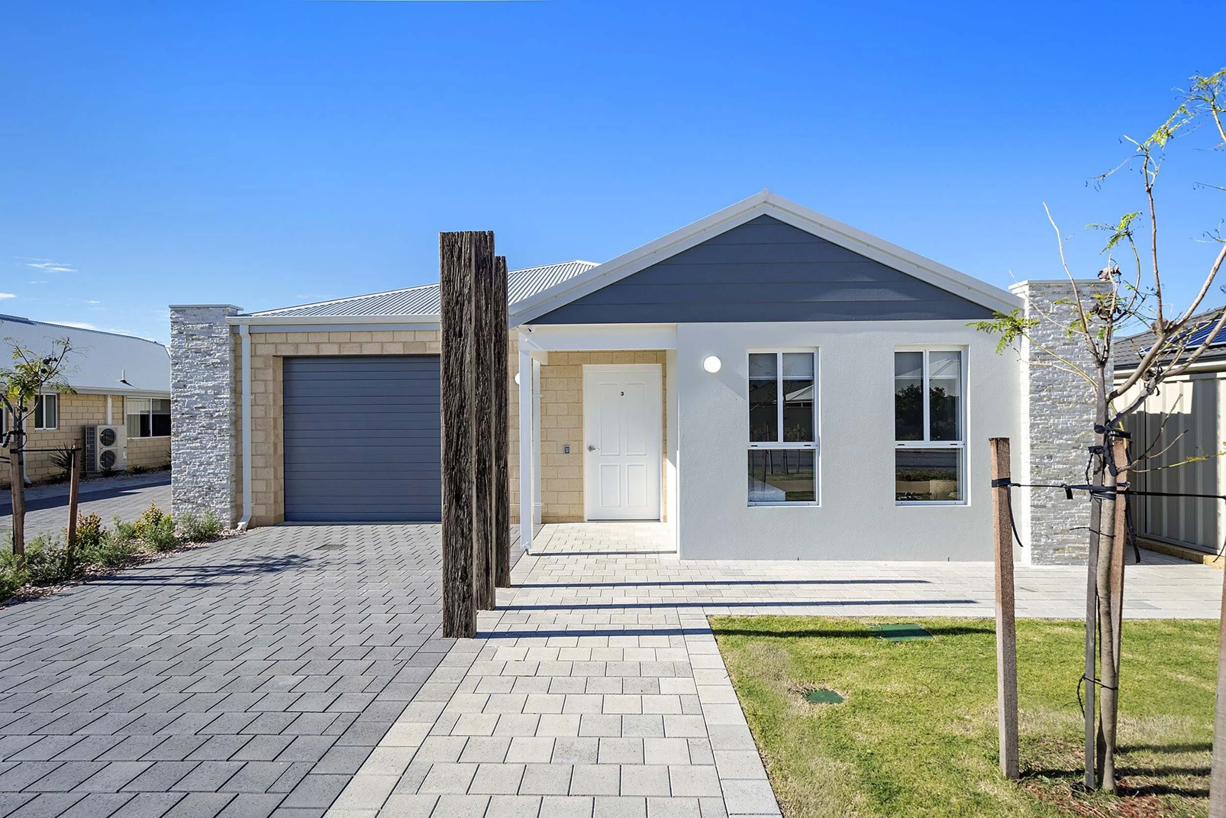 Modern single-story house with garage, gray roof, white and light gray exterior, paved driveway, and small front yard with young trees.
