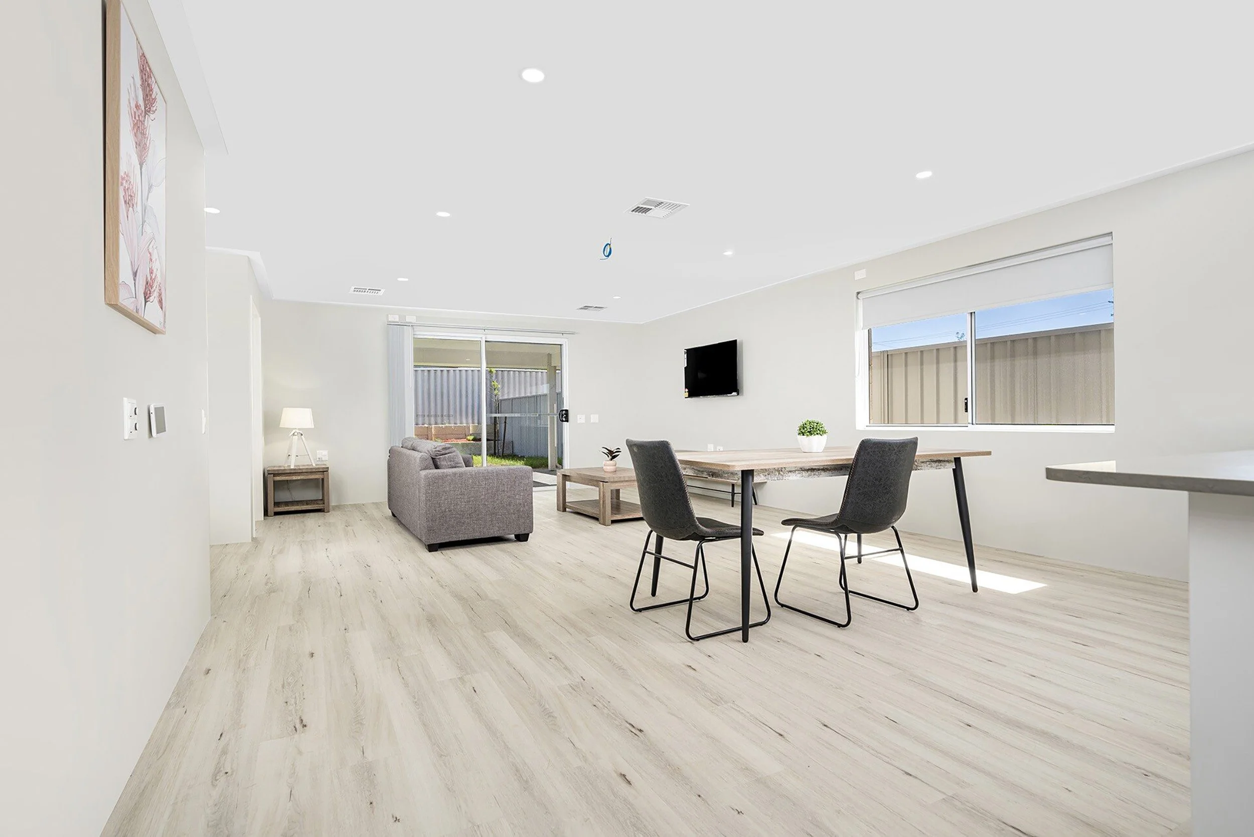 Modern open-plan living room with wooden flooring, a gray sofa, dining table with chairs, wall-mounted TV, window, and sliding glass doors leading to a backyard.