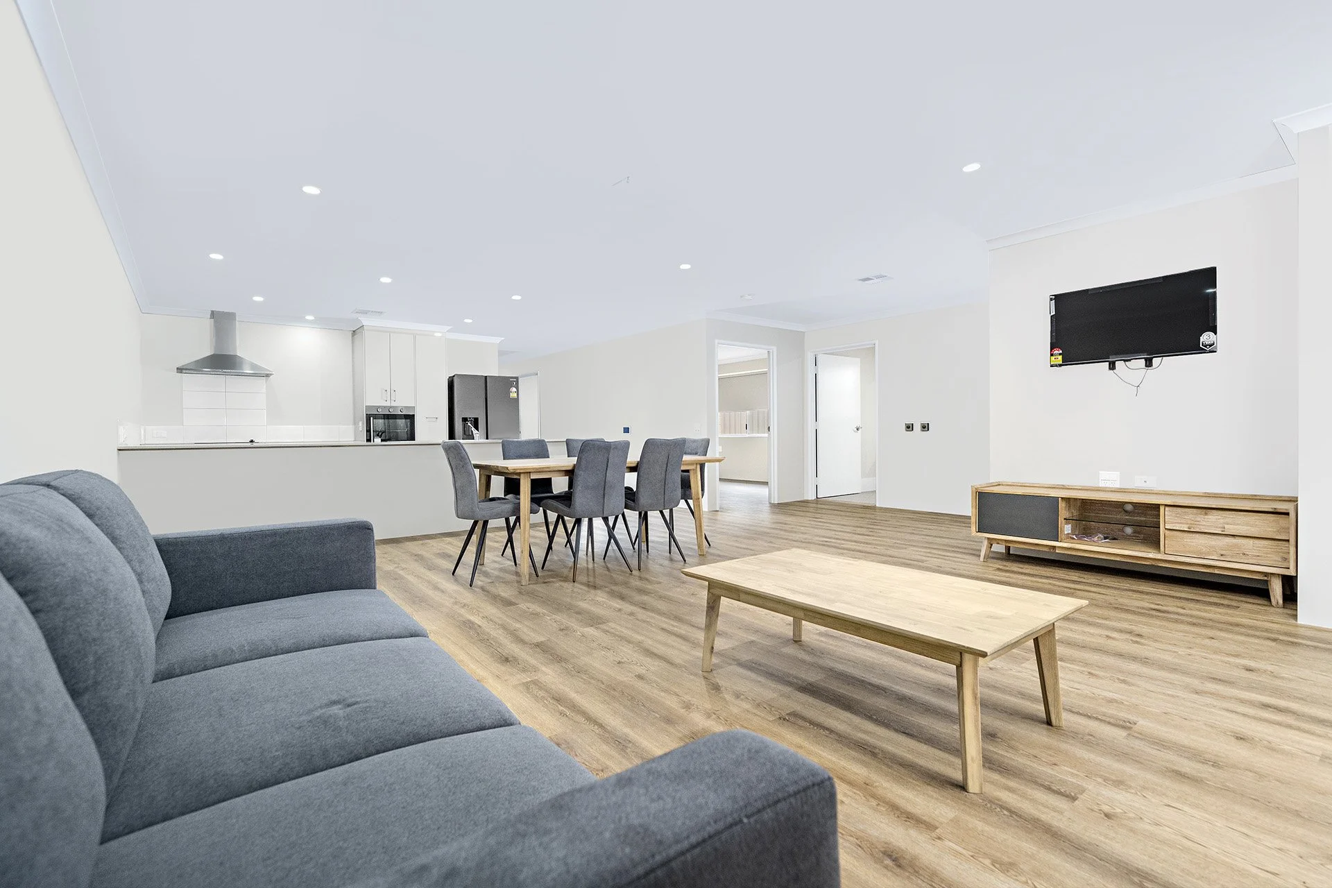 Modern open-plan living area with a gray sofa, wooden coffee table, dining table with chairs, and a kitchen in the background. Wall-mounted TV and wooden media console are visible.