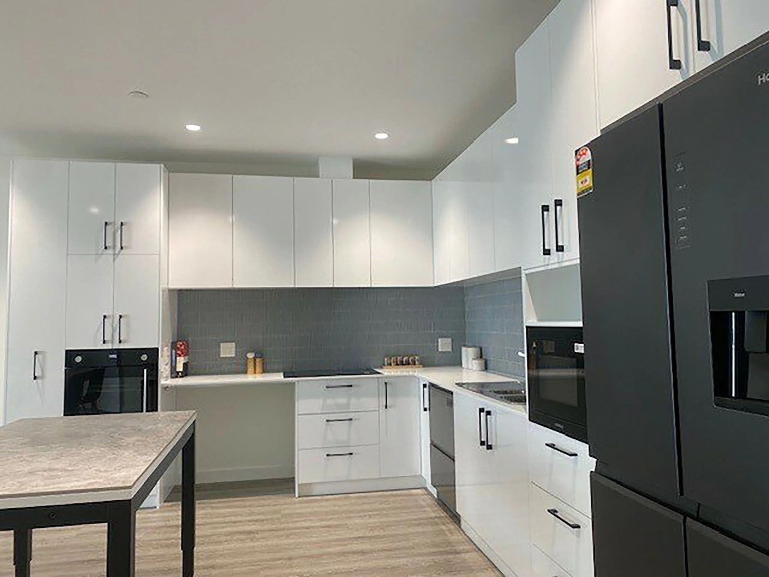 Modern kitchen with white cabinets, black appliances, and a gray marble countertop. Large refrigerator on the right, built-in oven on the left, and a tile backsplash.