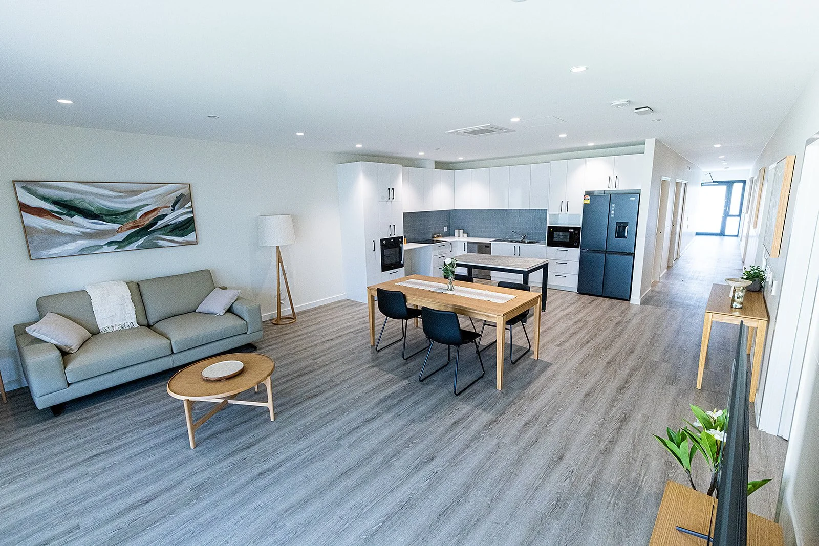 Modern open-plan living room and kitchen with gray sofa, wooden coffee table, dining table with chairs, white cabinetry, stainless steel appliances, and minimalistic decor.