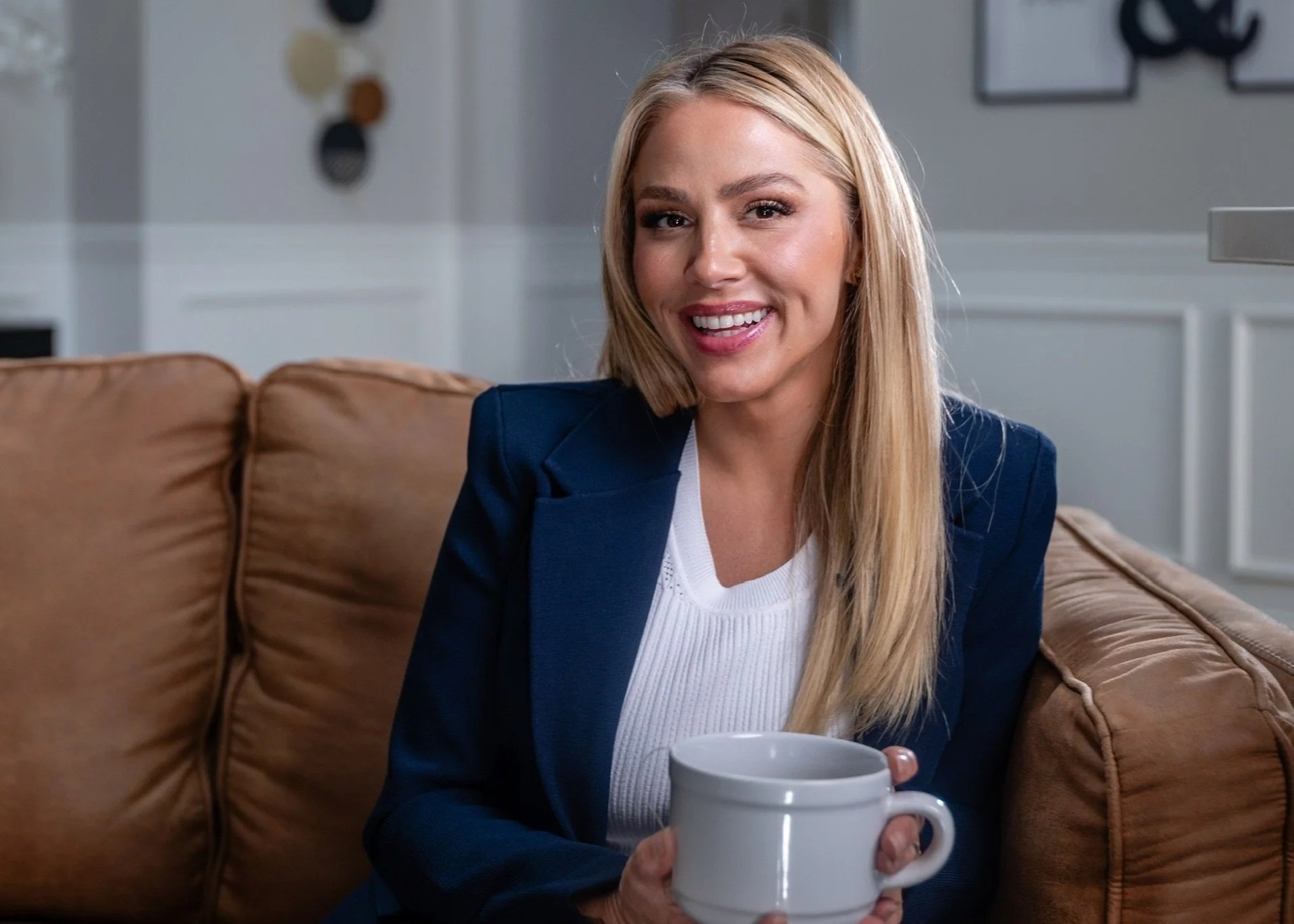 A smiling woman with long blonde hair sitting on a brown couch, holding a white mug, in a bright living room.