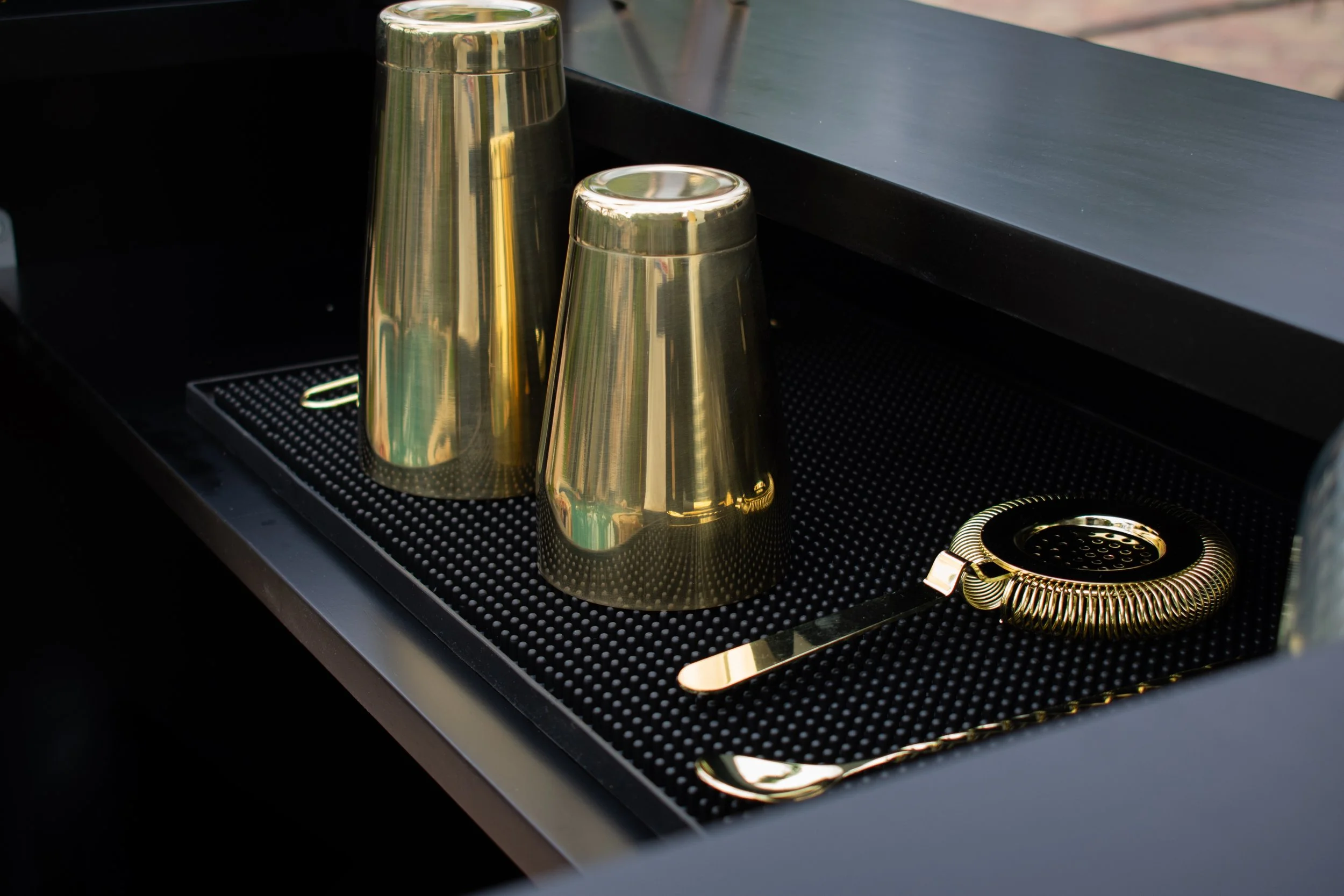 Golden cocktail shaker, two golden tumblers, and a gold bar spoon with a mesh strainer on a black bar mat.