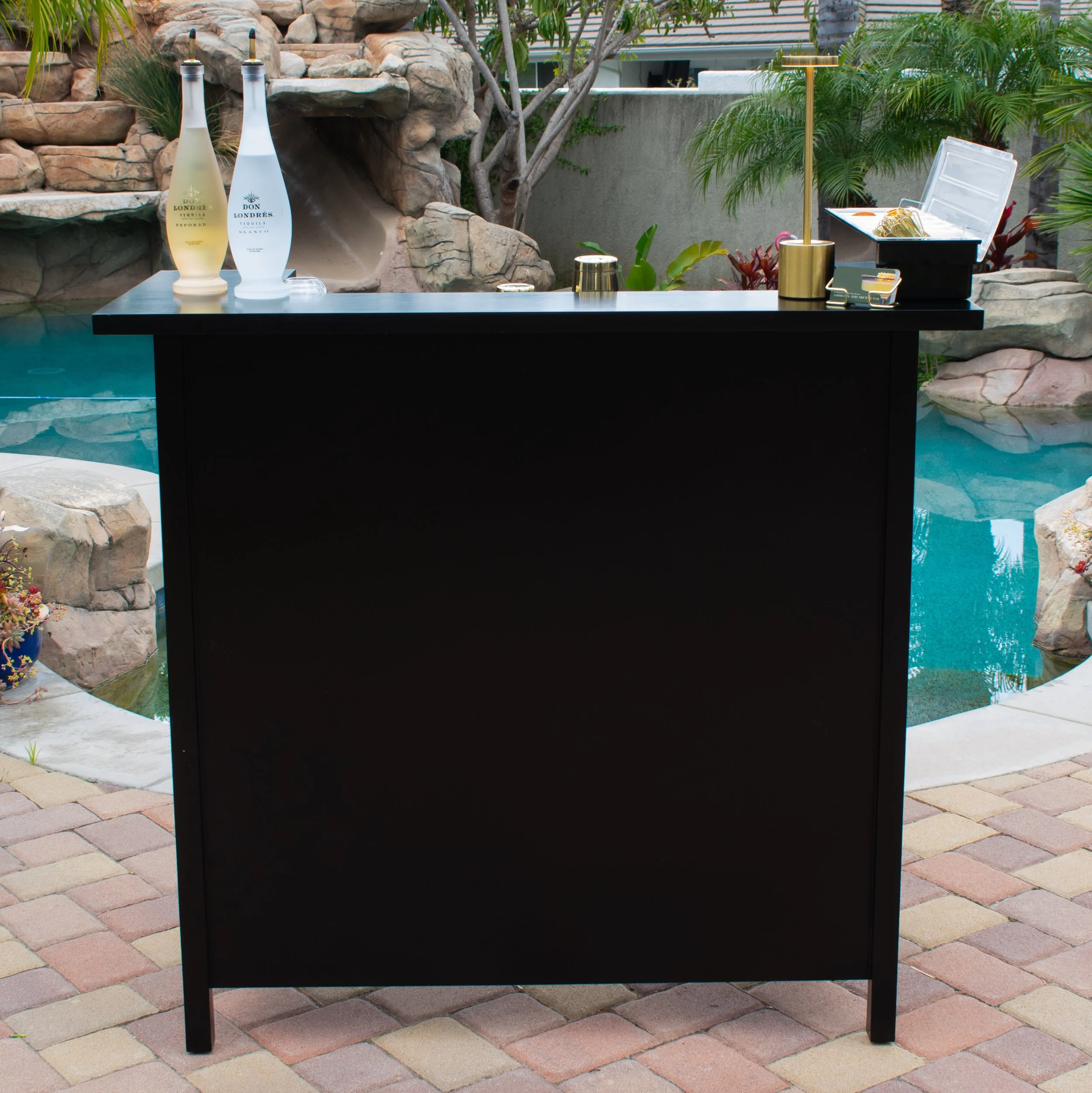 A black bar or counter with bottles, a small gold cup, and a lamp on it, set outdoors near a swimming pool with rocks and tropical plants in the background.