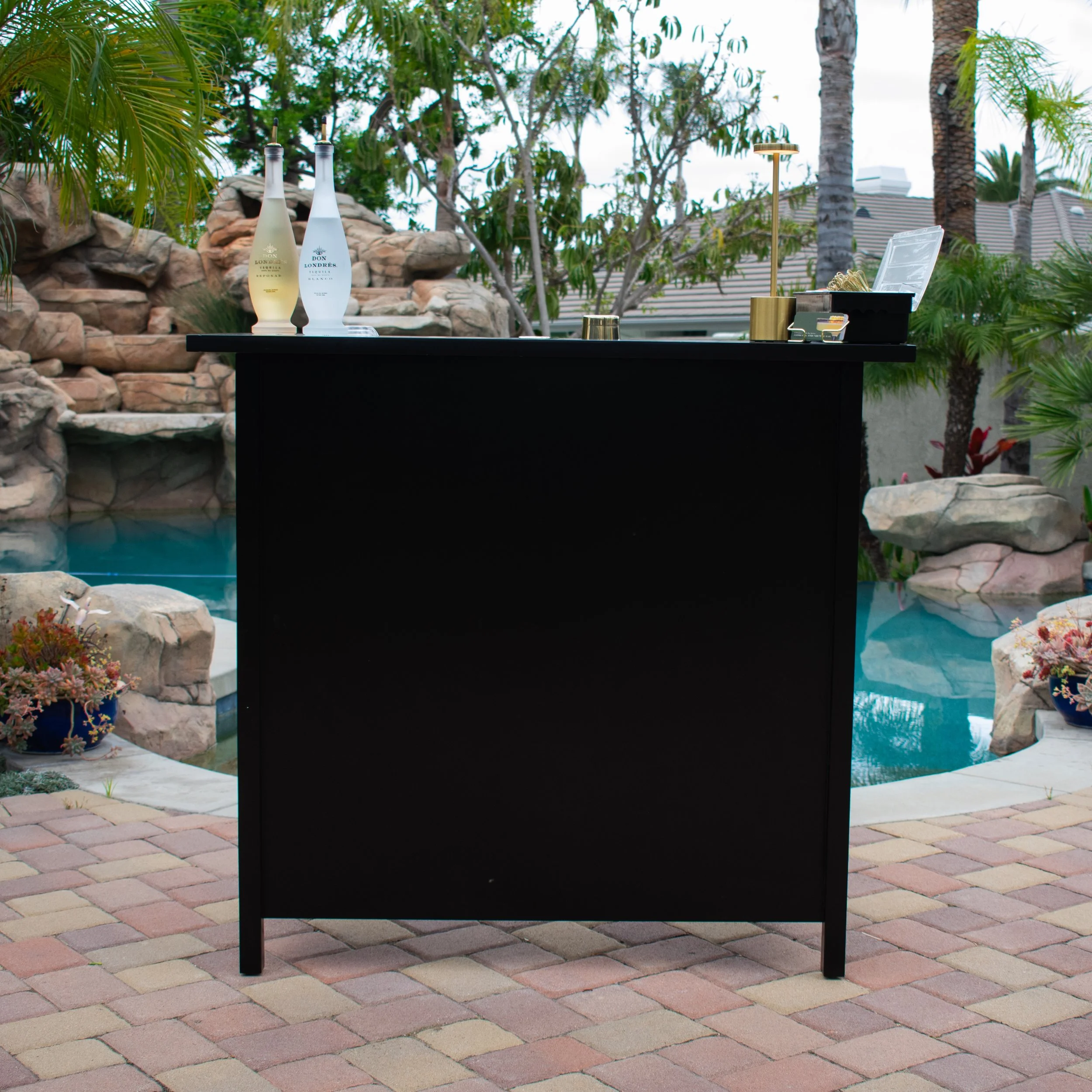 Outdoor bar setup with two bottles, a lamp, and a black counter, overlooking a pool and tropical plants.