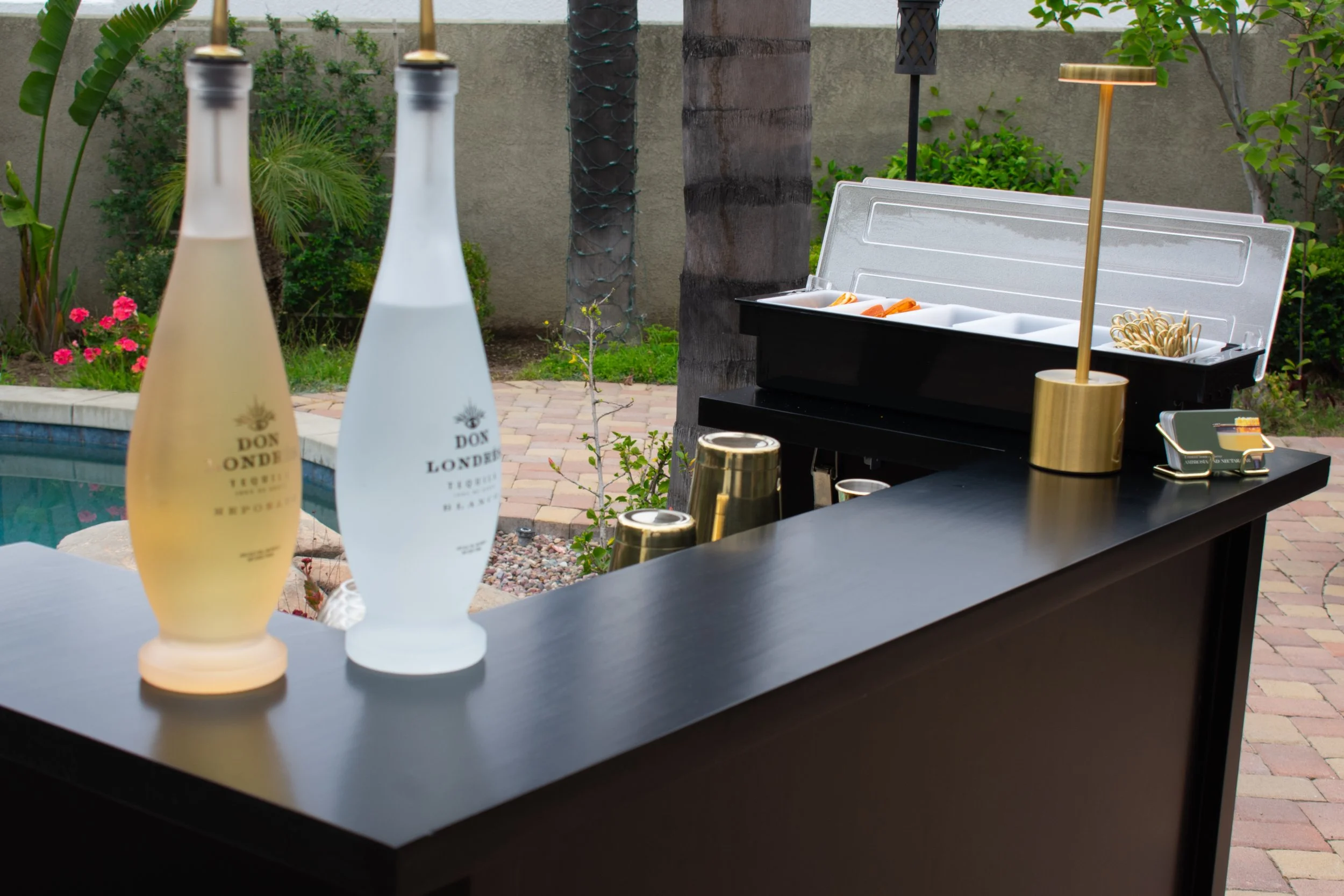 Outdoor bar setup with two bottles labeled Don London, a black food warmer, and black tables with gold accents, surrounded by a garden with plants and flowers.