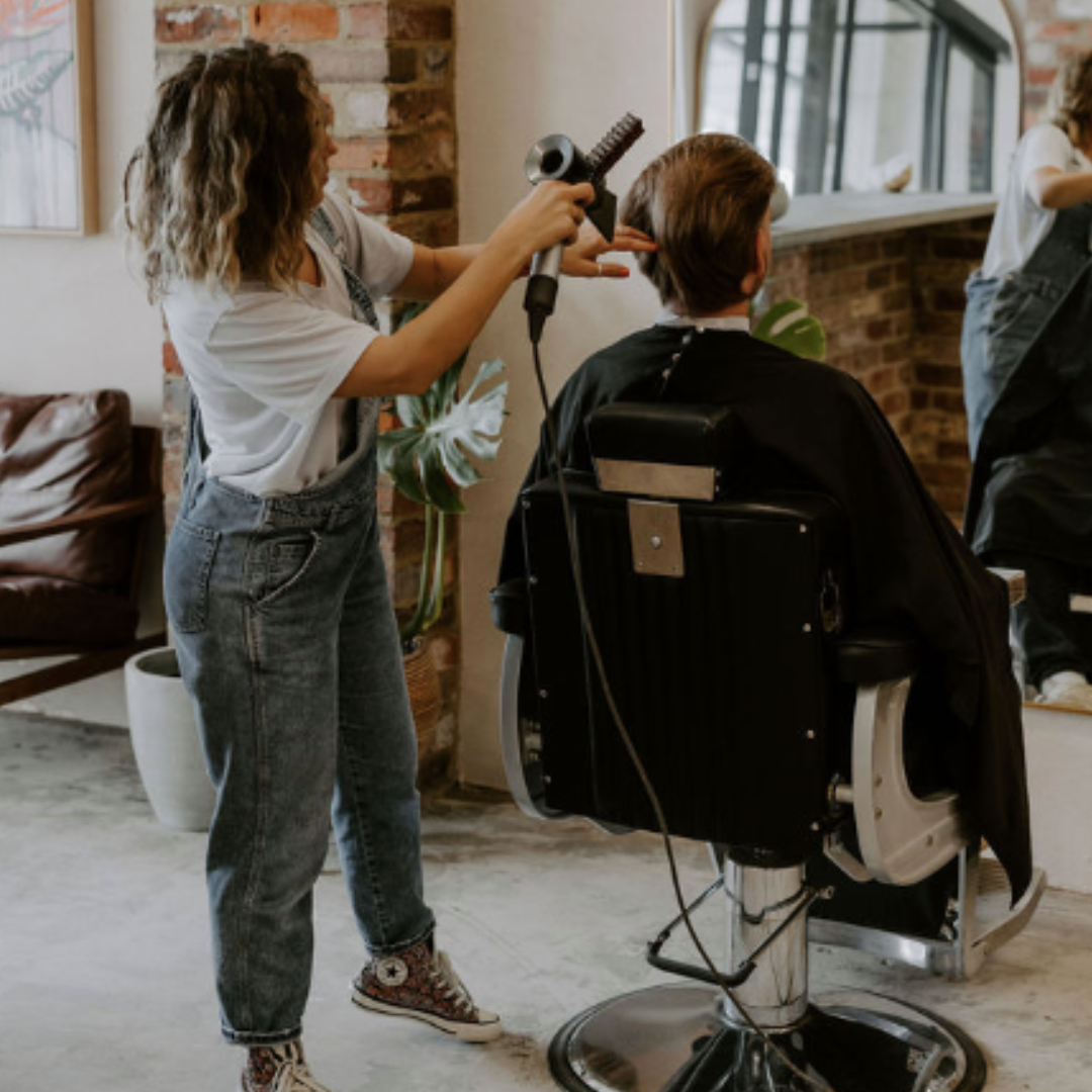 A woman styling a man's hair at a barbershop or salon, with a hairdryer in hand and a cape around the man.