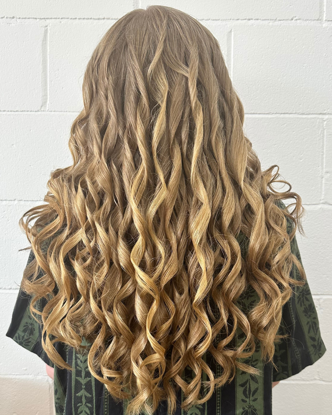Back view of a woman with long, blonde, curly hair against a white brick wall.