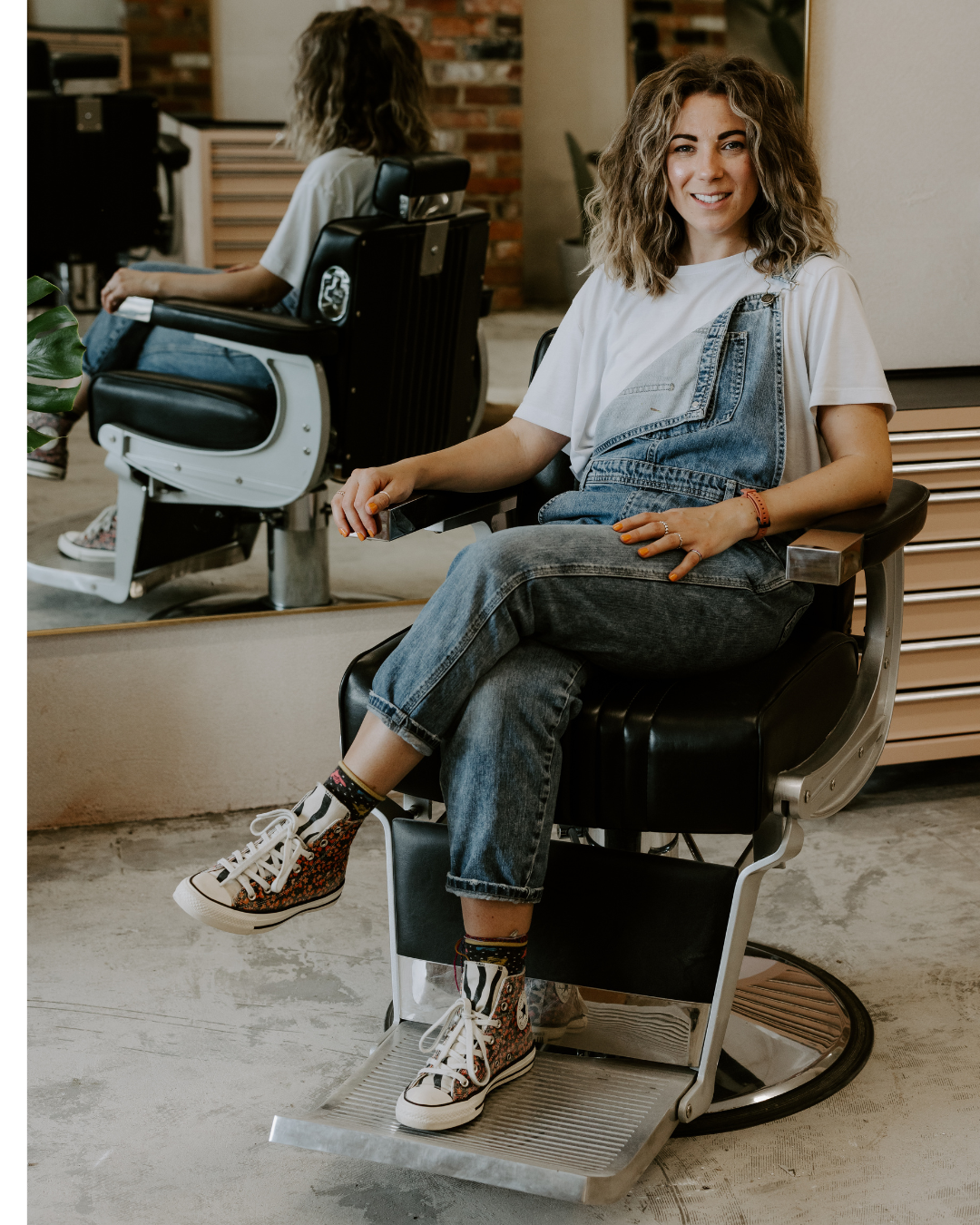 A woman sitting in a barber's chair at a salon, smiling, with curly hair, wearing a white t-shirt, denim overalls, colorful patterned sneakers, and socks.