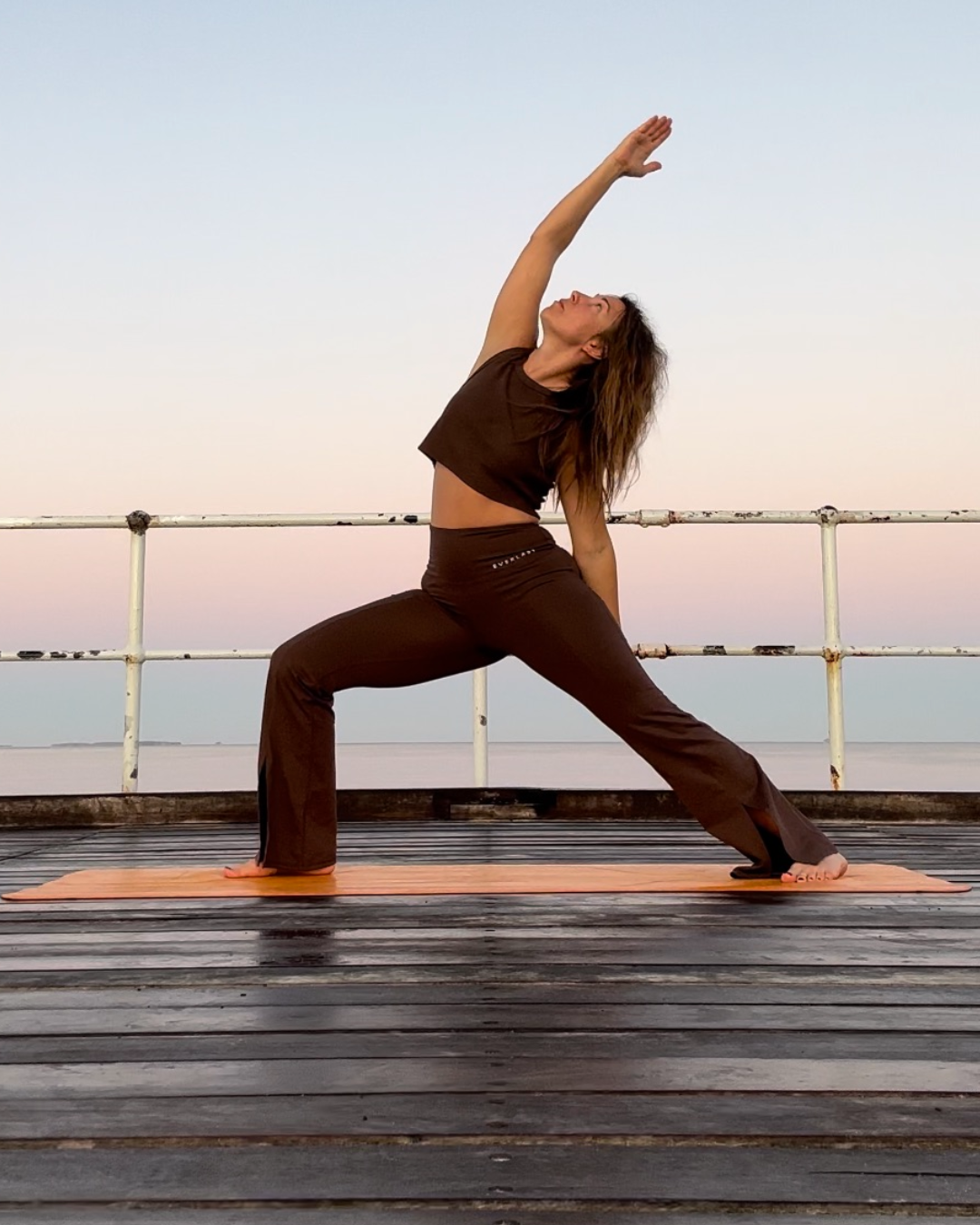 A woman practicing yoga outdoors on a wooden deck during sunset or sunrise, performing a side stretch pose in front of a calm body of water with a pastel sky.