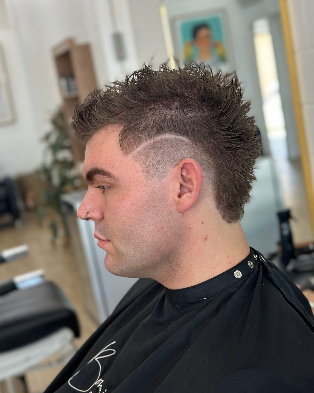 Side profile of a young man with a mohawk hairstyle featuring a shaved design on the side, sitting in a hair salon.