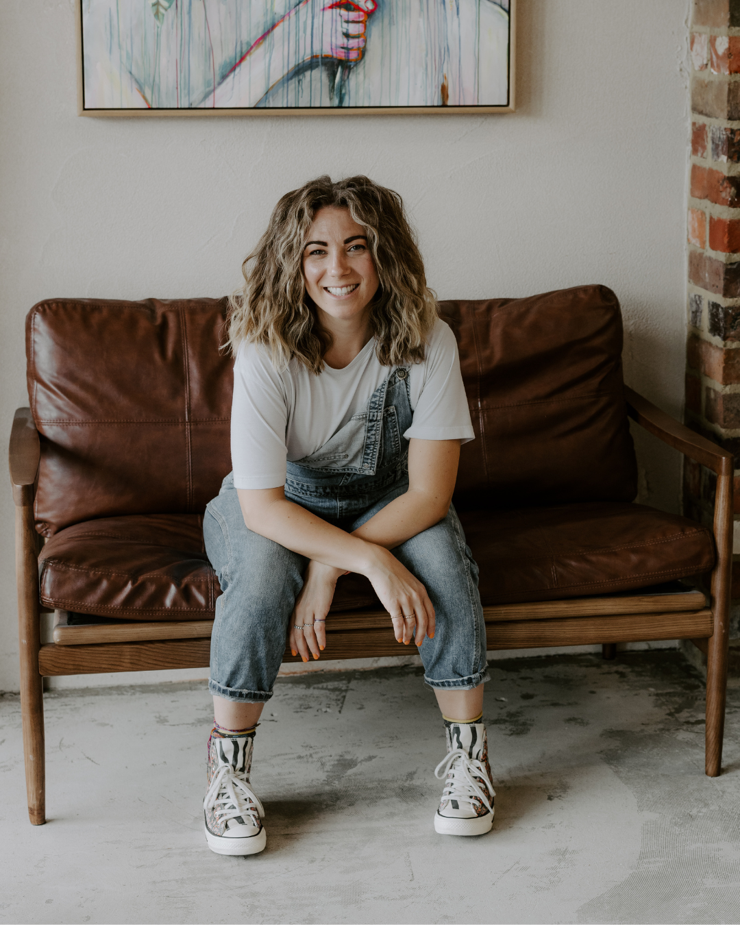 A woman with curly hair smiling at the camera, sitting on a brown leather couch. She is wearing a white t-shirt, denim overalls, and zebra-striped sneakers, with a colorful bracelet on her ankle. She is in a room with a concrete floor, a partially exposed brick wall, and a framed abstract artwork hanging behind her.