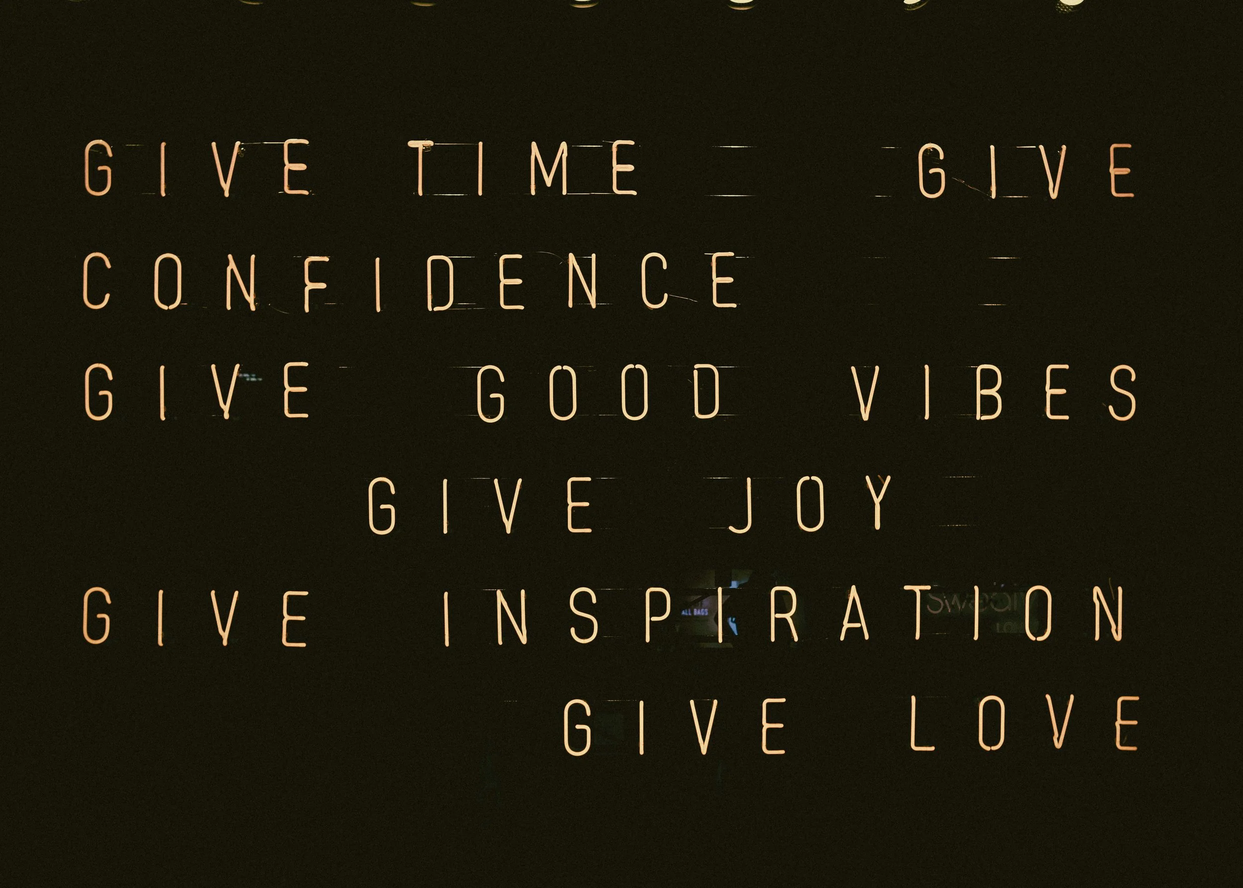 A dark wall with illuminated text displaying inspirational phrases: "GIVE TIME GIVE CONFIDENCE GIVE GOOD VIBES GIVE JOY GIVE INSPIRATION GIVE LOVE."