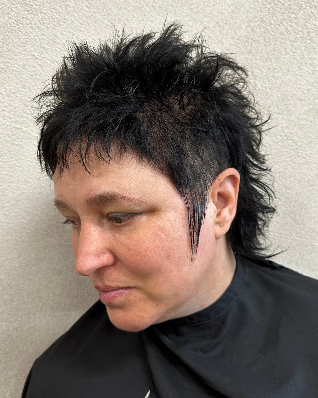 A woman with short, black, spiky and layered haircut facing downward against a plain wall.