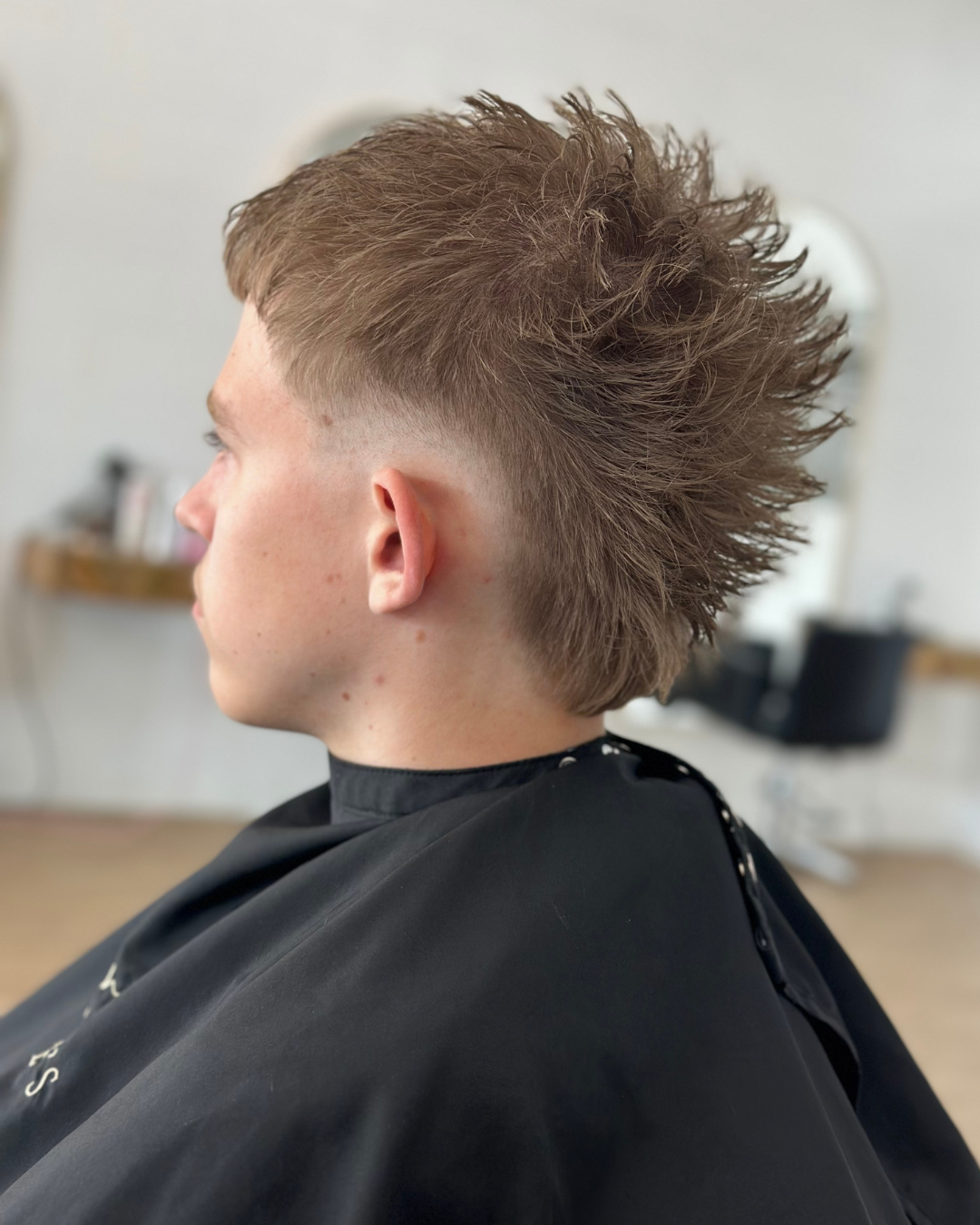 Young man with a mohawk hairstyle in a barbershop, seated in a barber's cape, with a blurred background.
