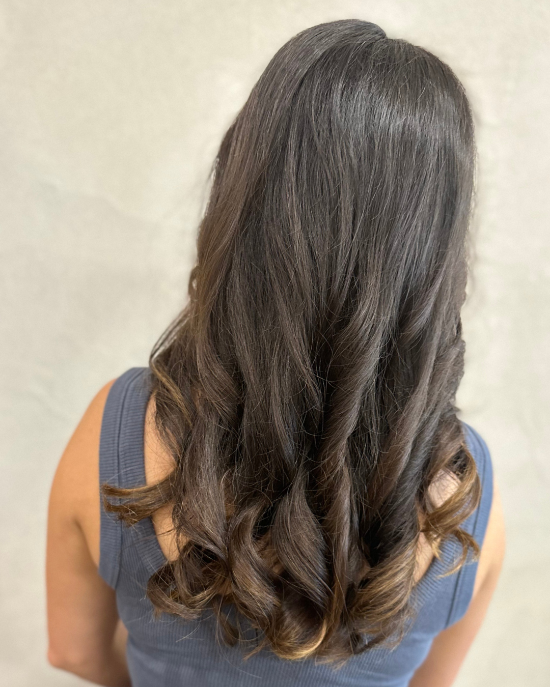 Back view of a woman with long, wavy brunette hair wearing a sleeveless blue top.