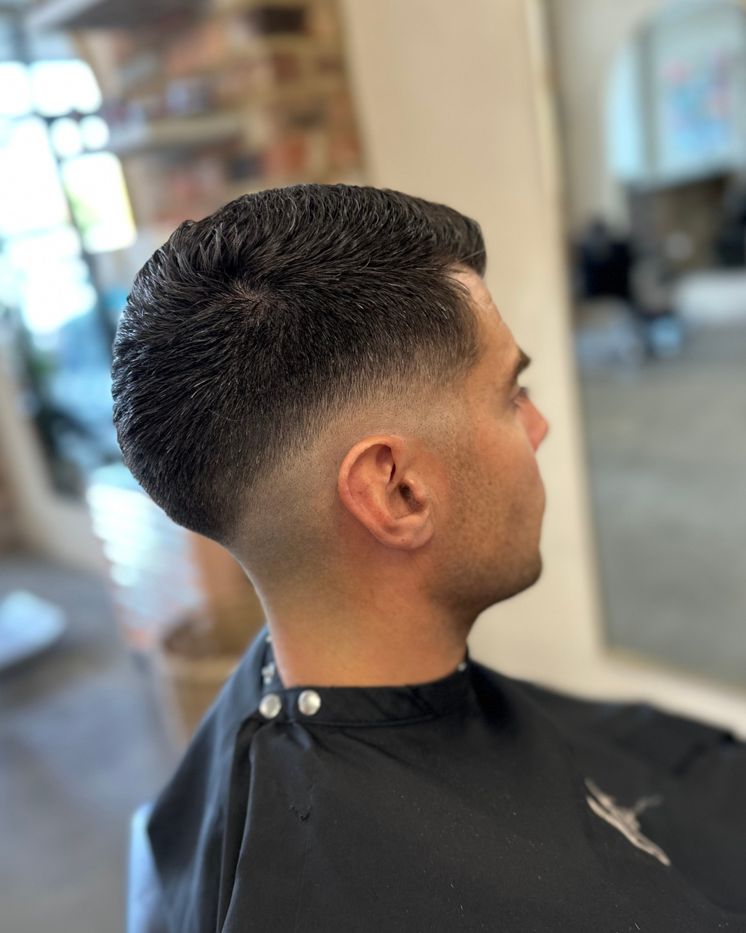 A man with a fresh fade haircut sitting in a barbershop, facing to the right, with a black cape over his shoulders.